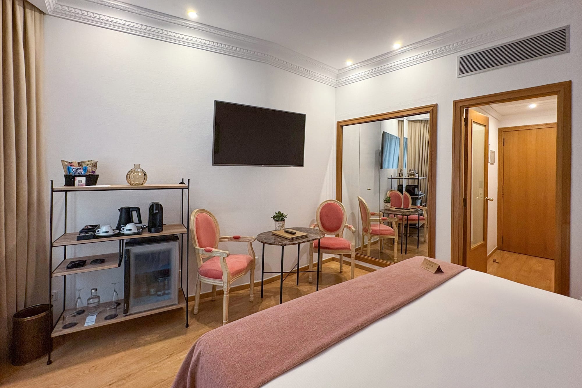 Modern hotel room with bed, minibar, wall-mounted TV, pink chairs, and large mirror.