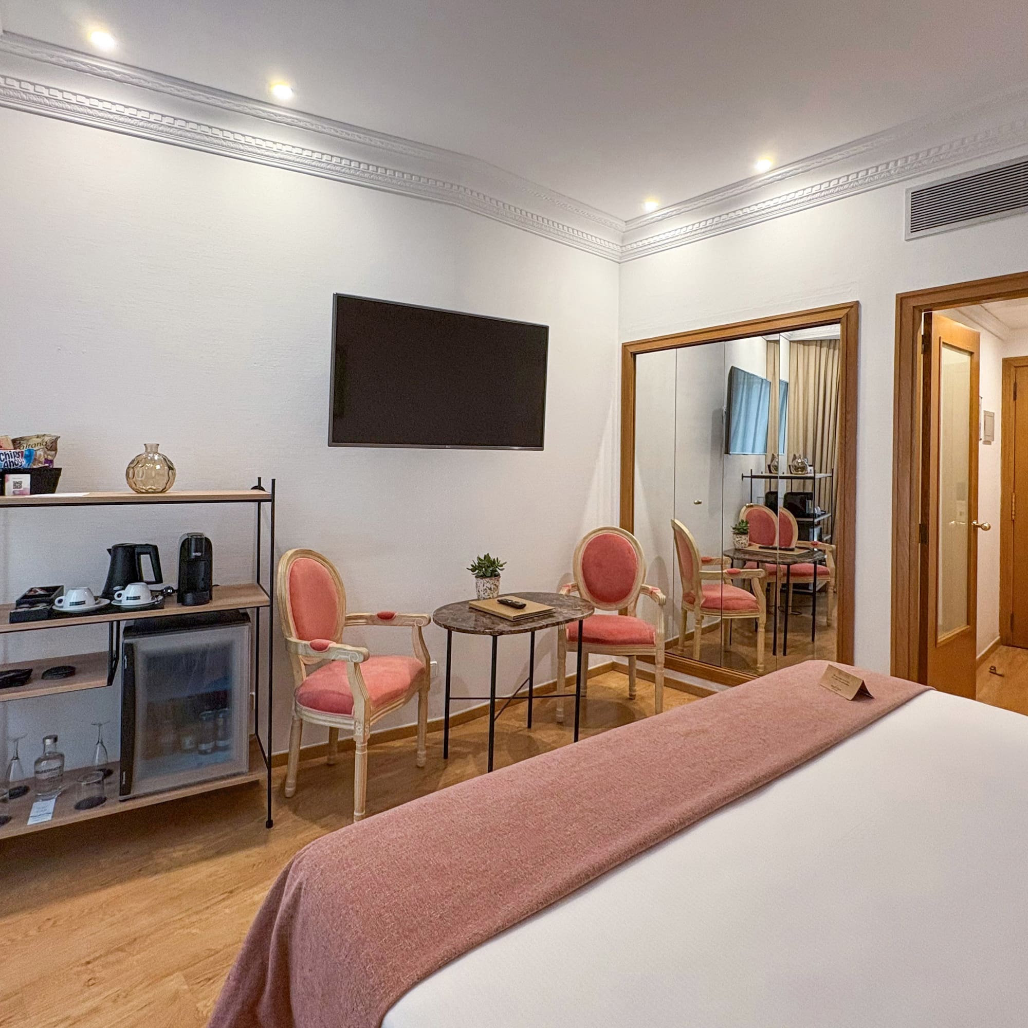 Modern hotel room with bed, minibar, wall-mounted TV, pink chairs, and large mirror.