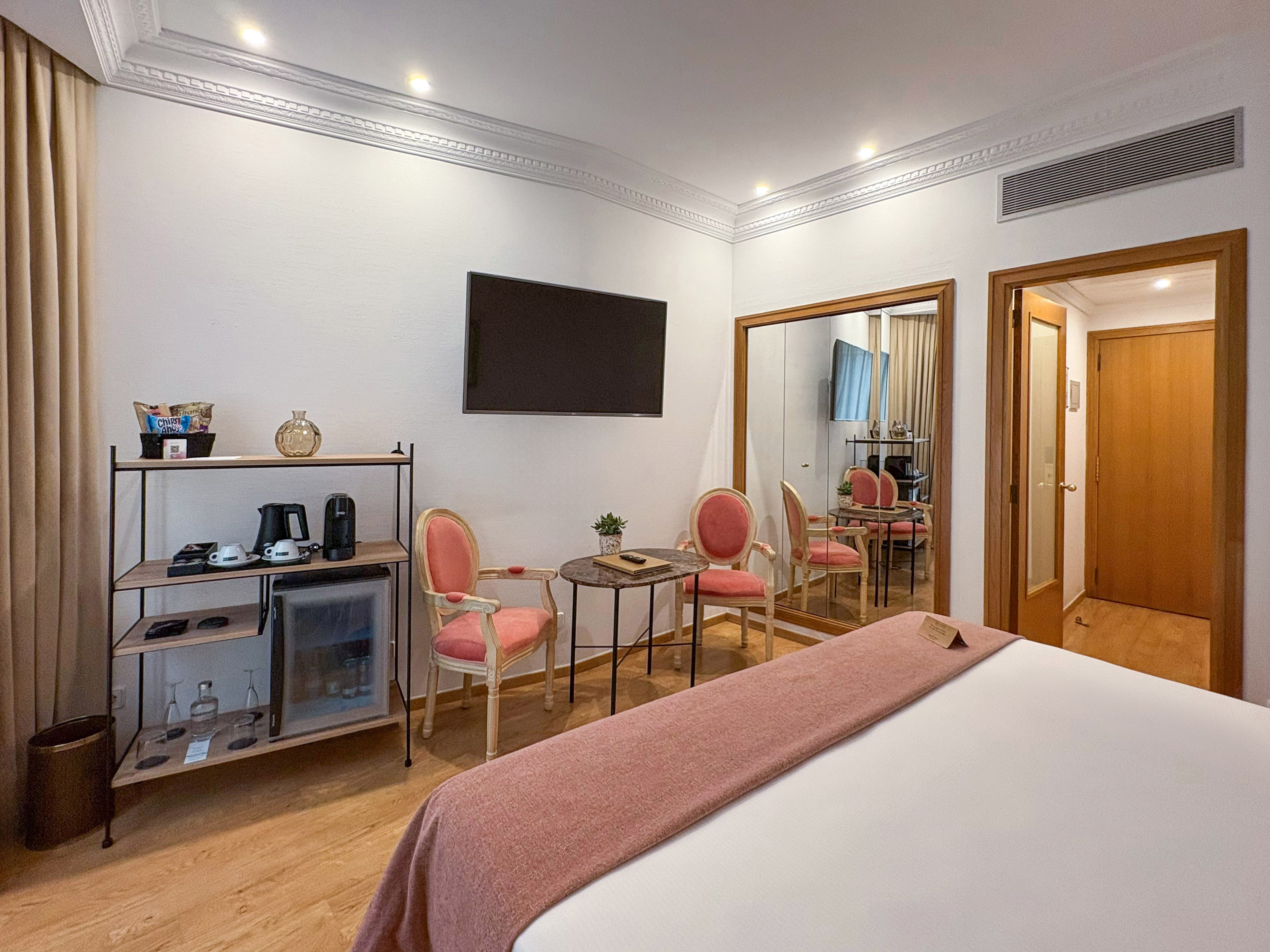 Modern hotel room with bed, minibar, wall-mounted TV, pink chairs, and large mirror.