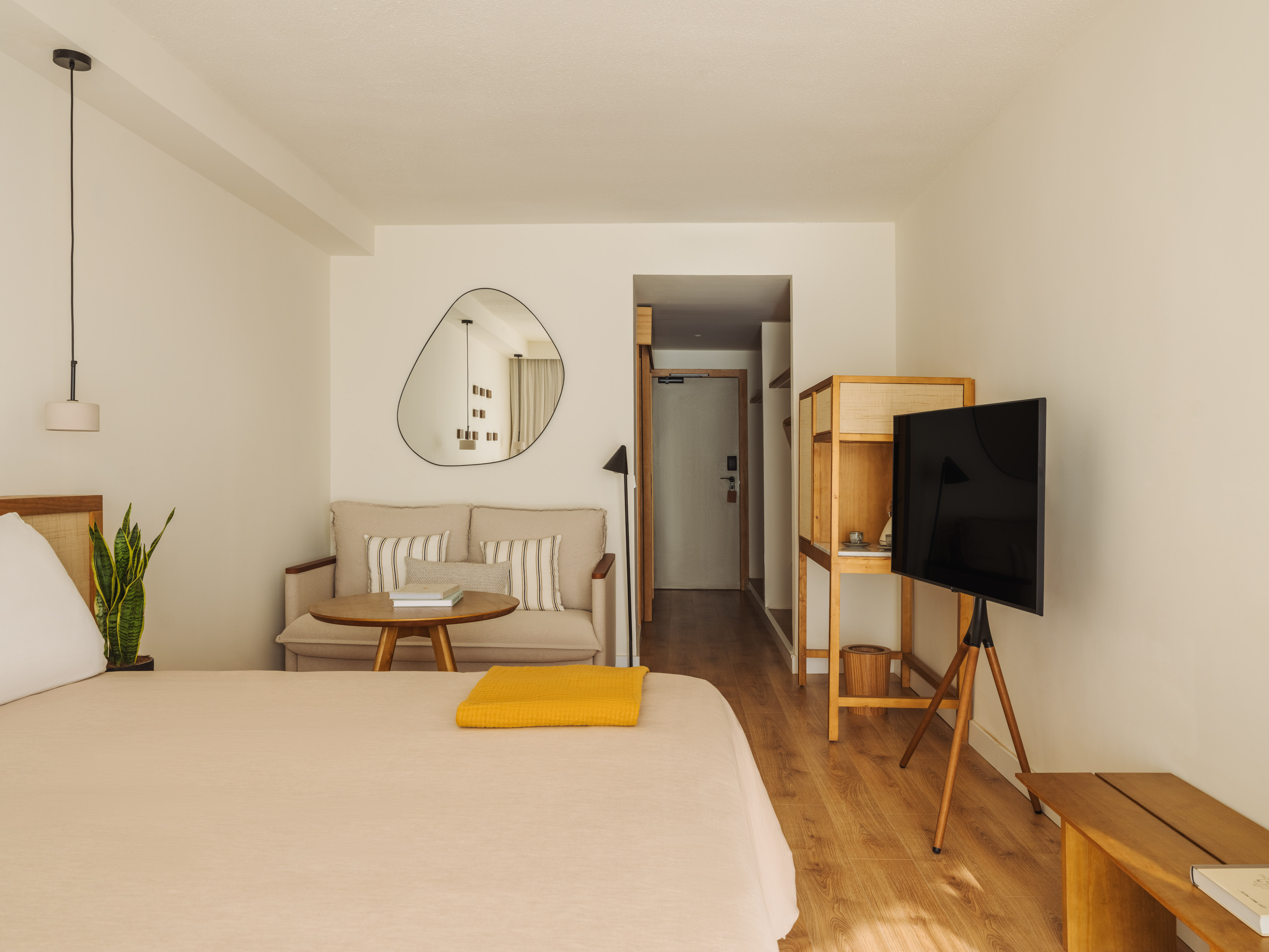 Minimalist hotel room with bed, neutral sofa, mirror, and modern tripod TV.