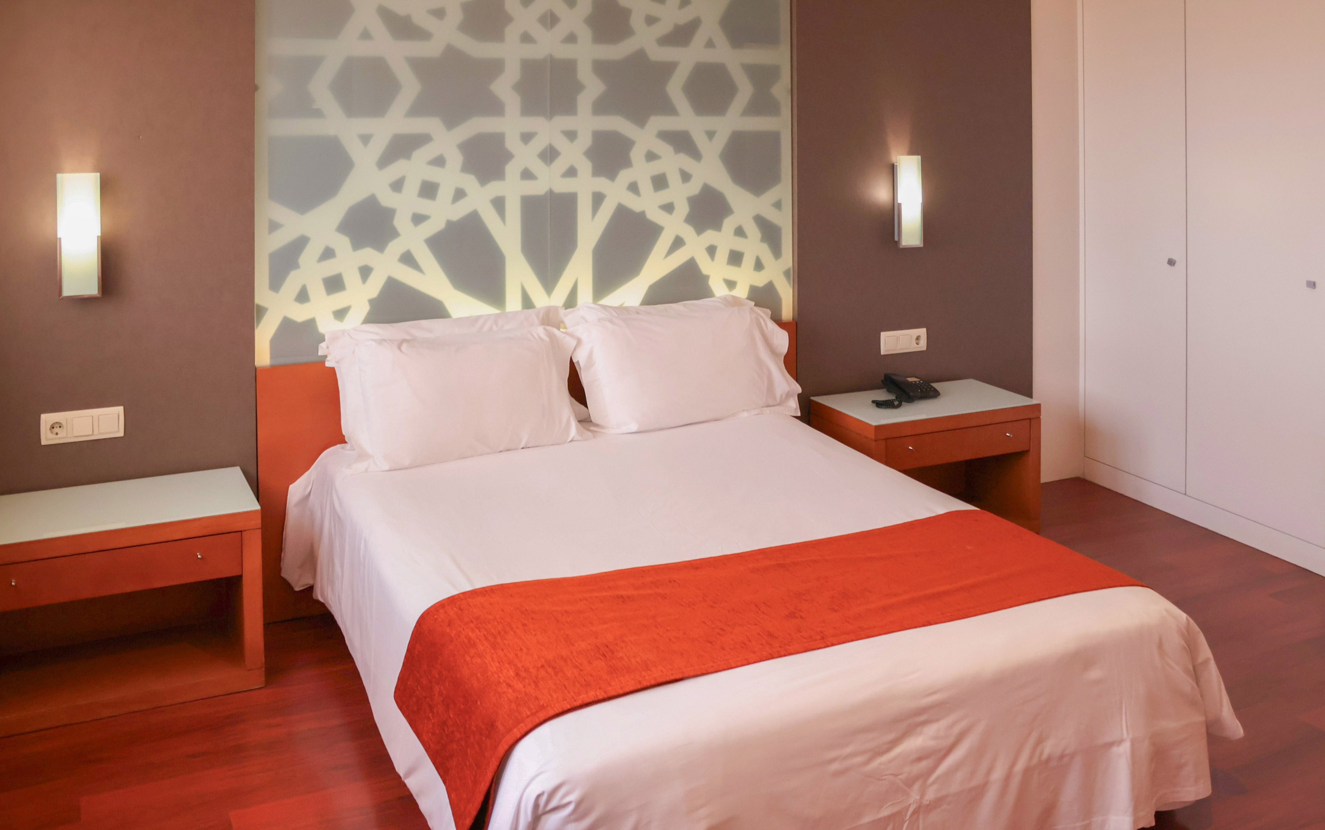 Modern hotel room with double bed, orange runner, and geometric pattern headboard.