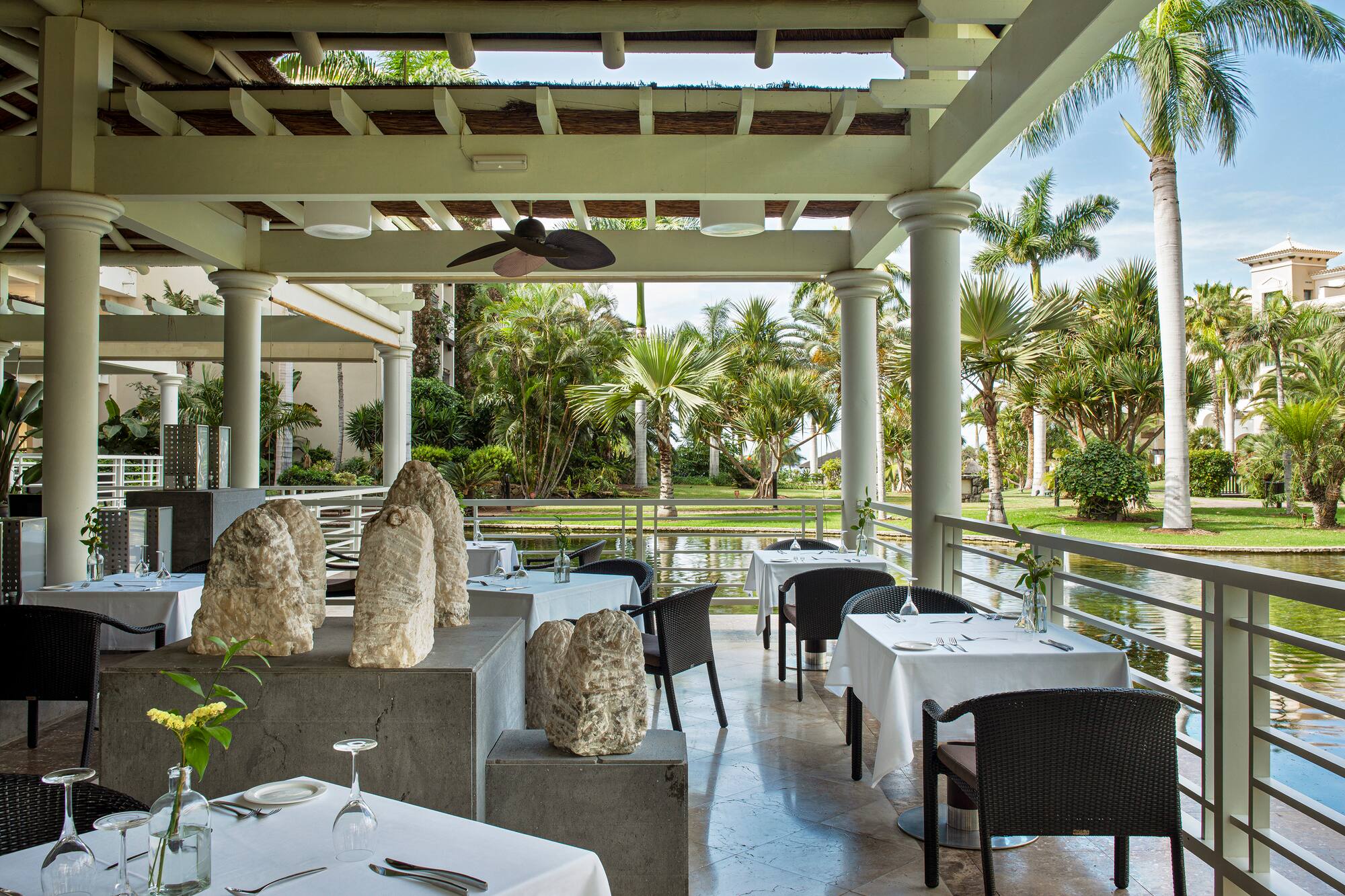 Tropical outdoor restaurant patio with set tables, lush garden, and water view.