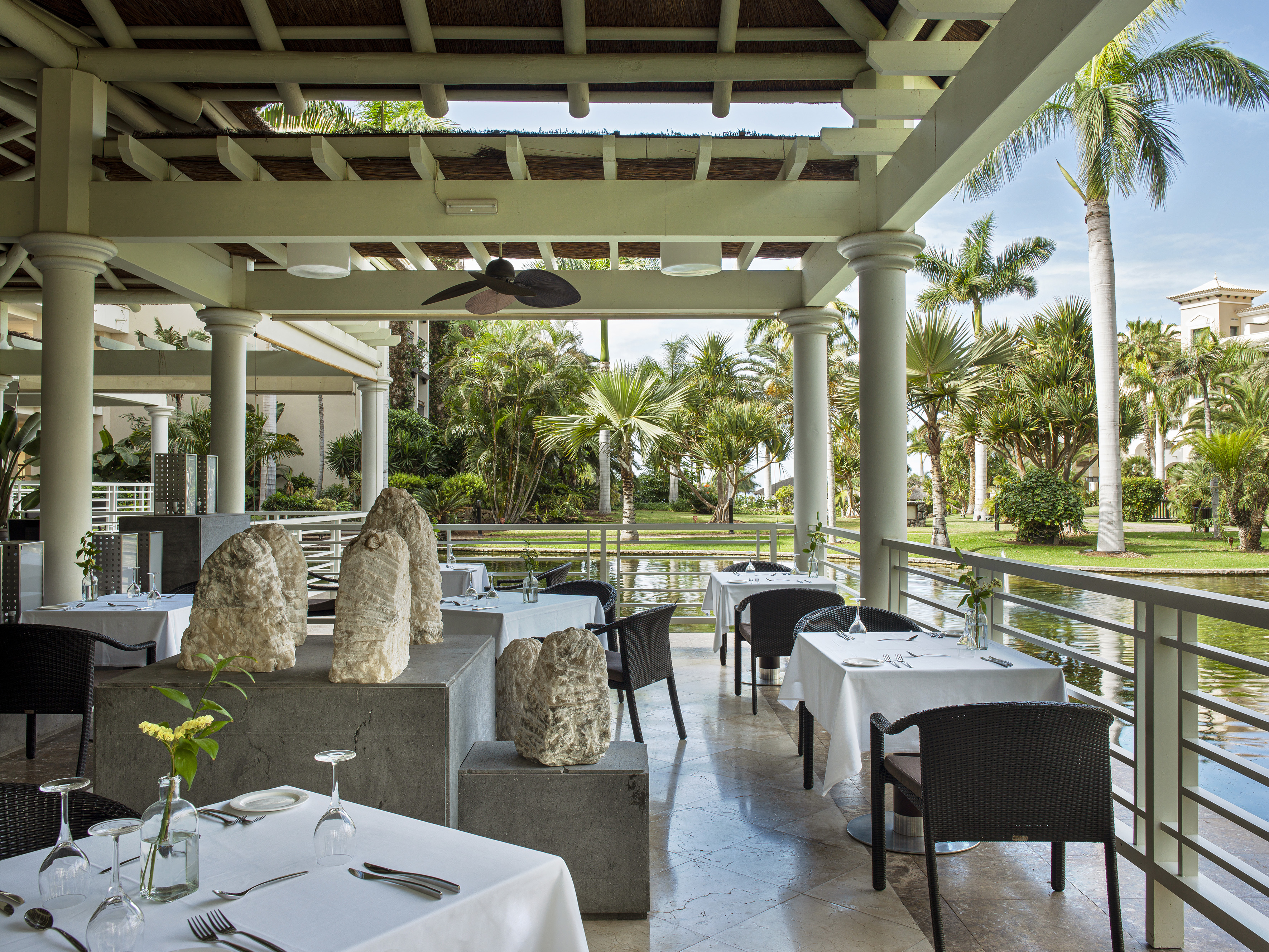 Tropical outdoor restaurant patio with set tables, lush garden, and water view.