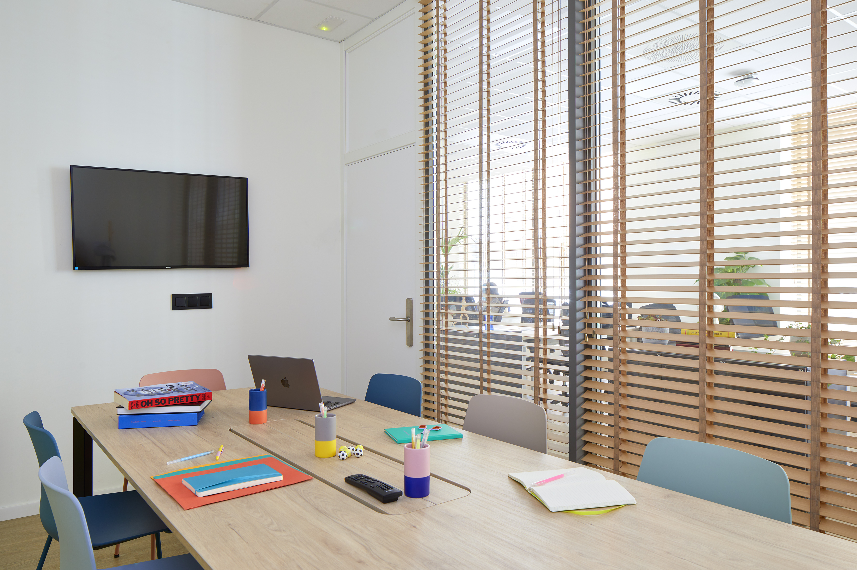 Modern office meeting room with wooden table, colorful chairs, TV screen, and blinds.