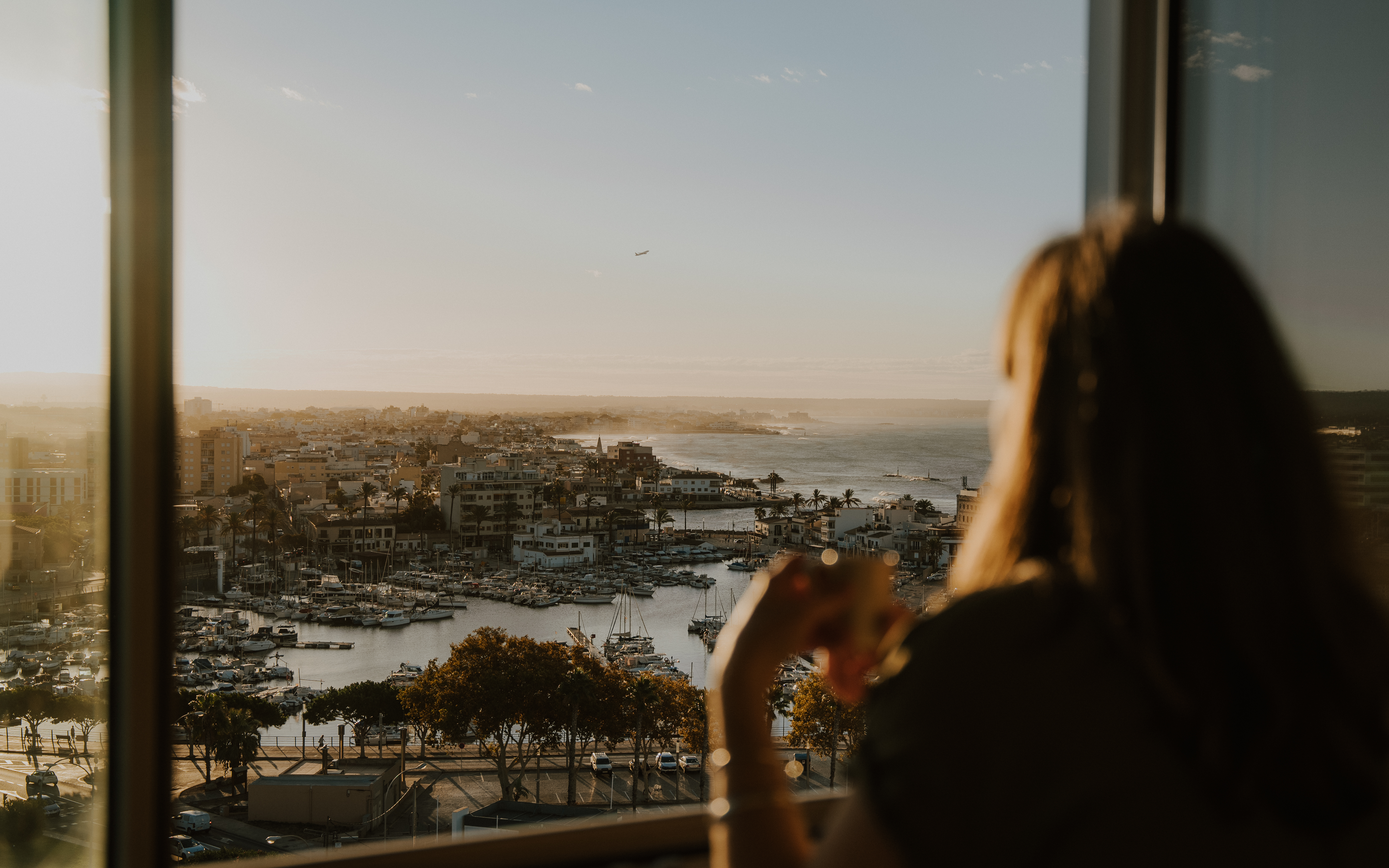 Mujer viendo puerto, ciudad costera y mar al atardecer desde ventana.