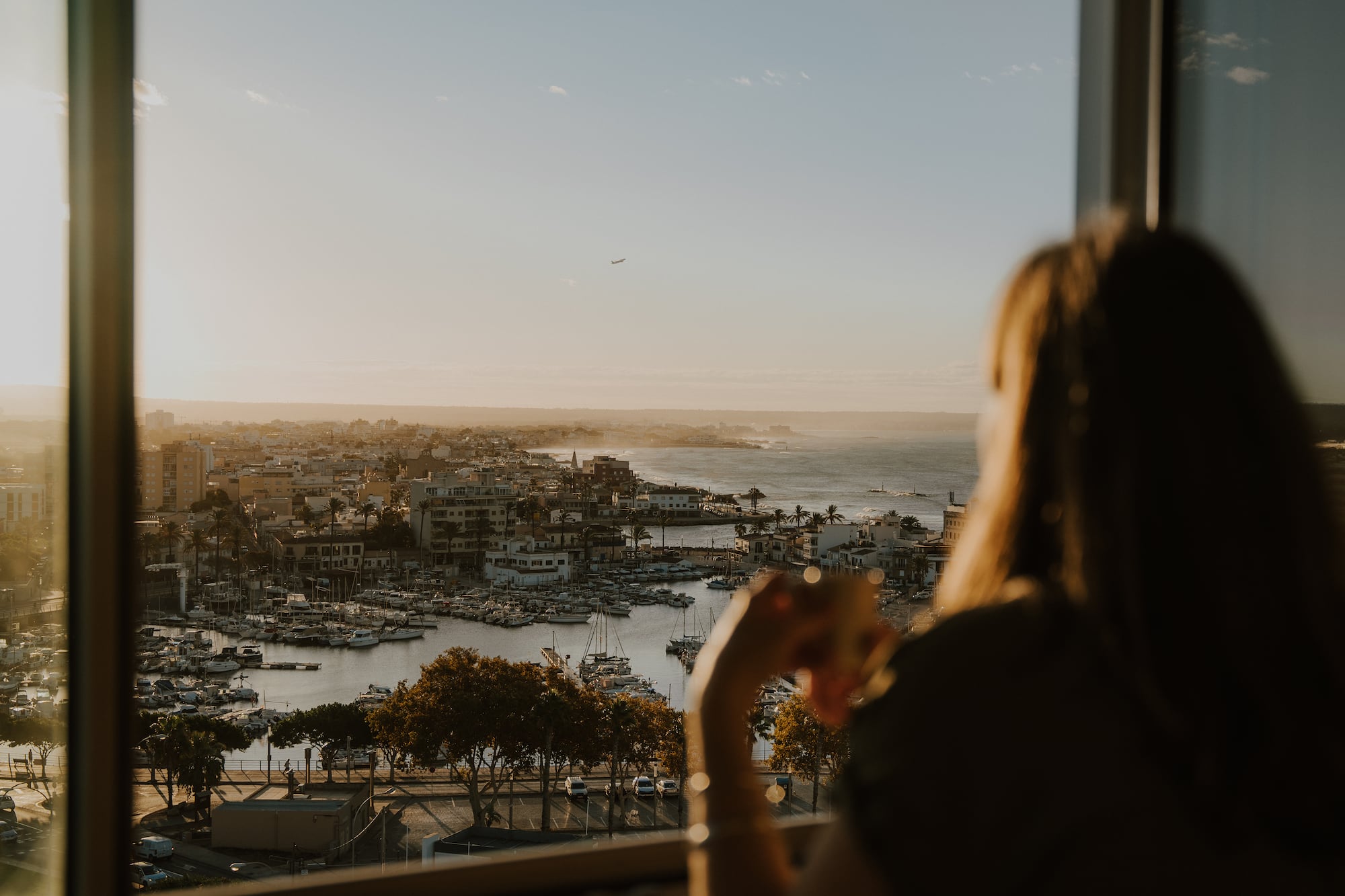 Mujer viendo puerto, ciudad costera y mar al atardecer desde ventana.
