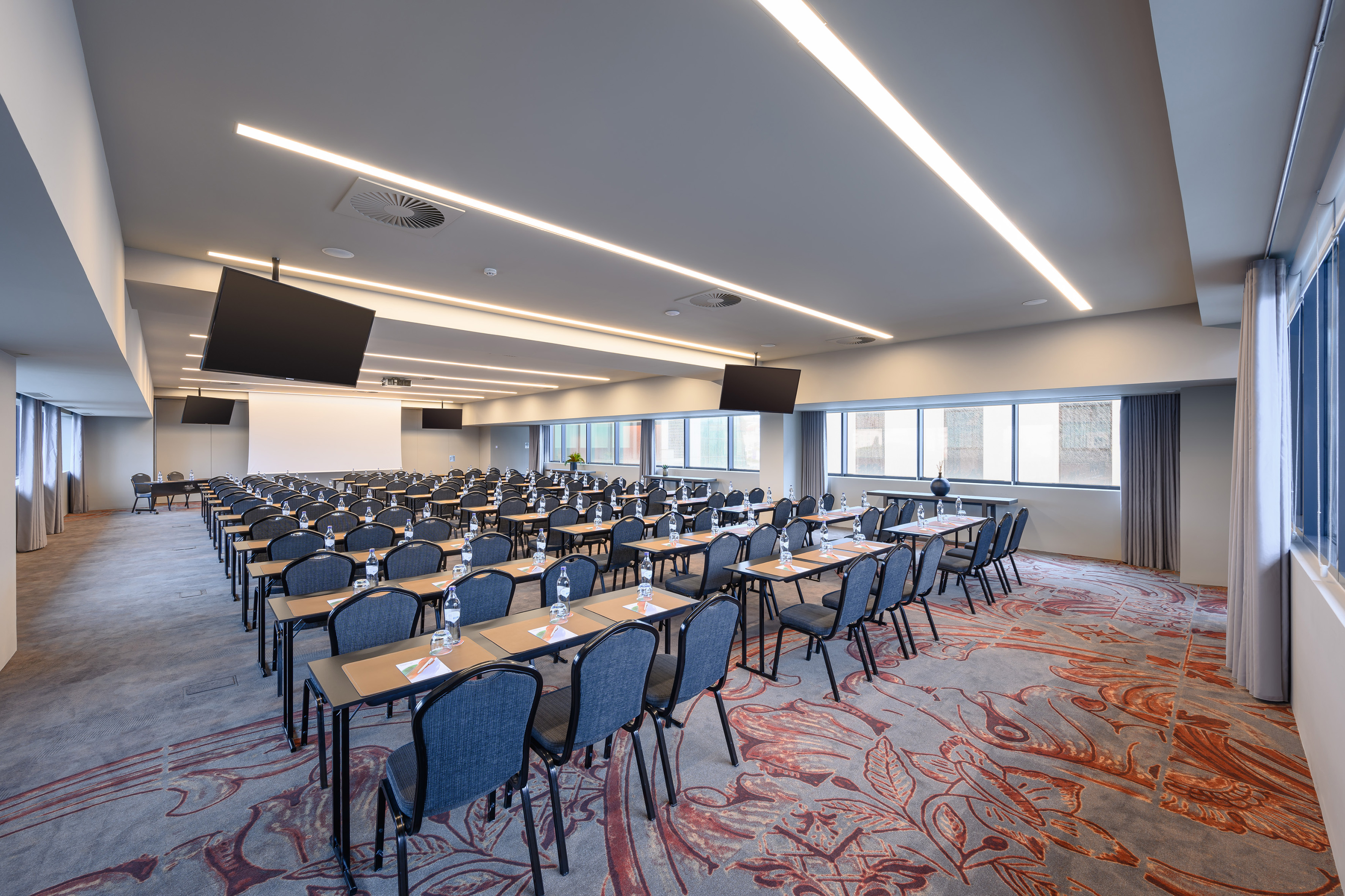 Modern conference room with rows of tables, chairs, and multiple screens.