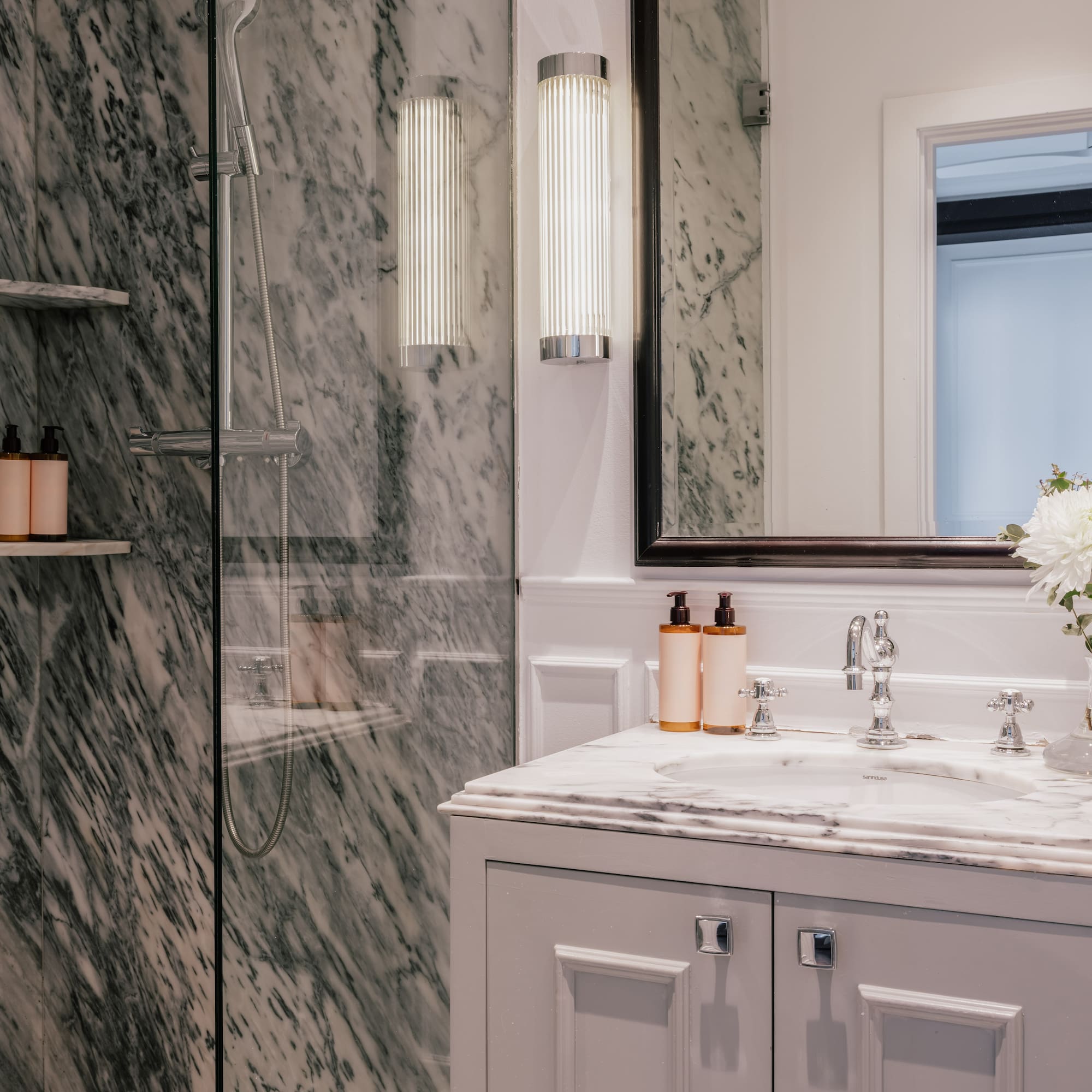 Luxury marble bathroom featuring glass shower, white vanity, mirror, and chrome details.
