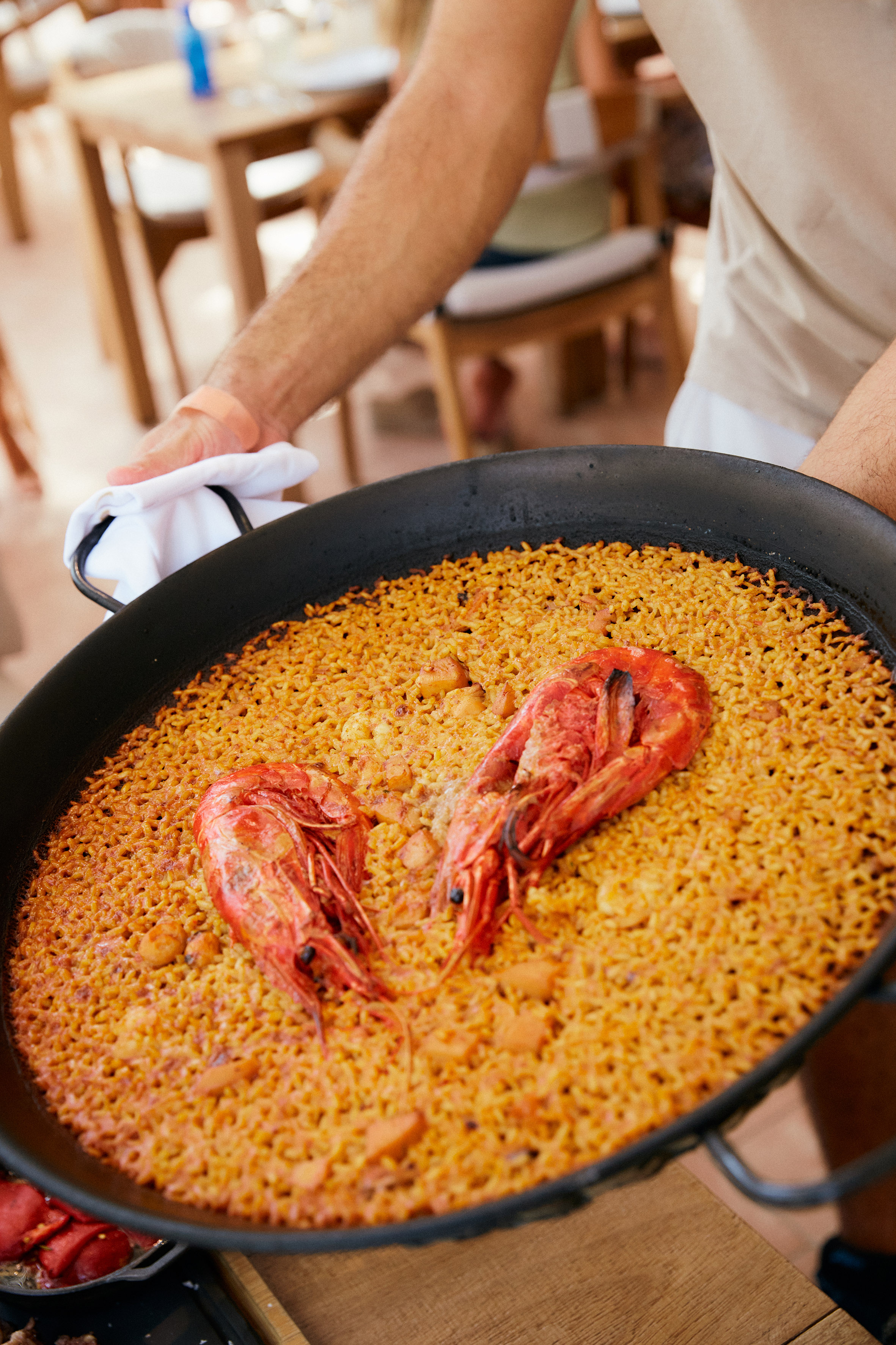 Hands serving large seafood paella with rice and red prawns.