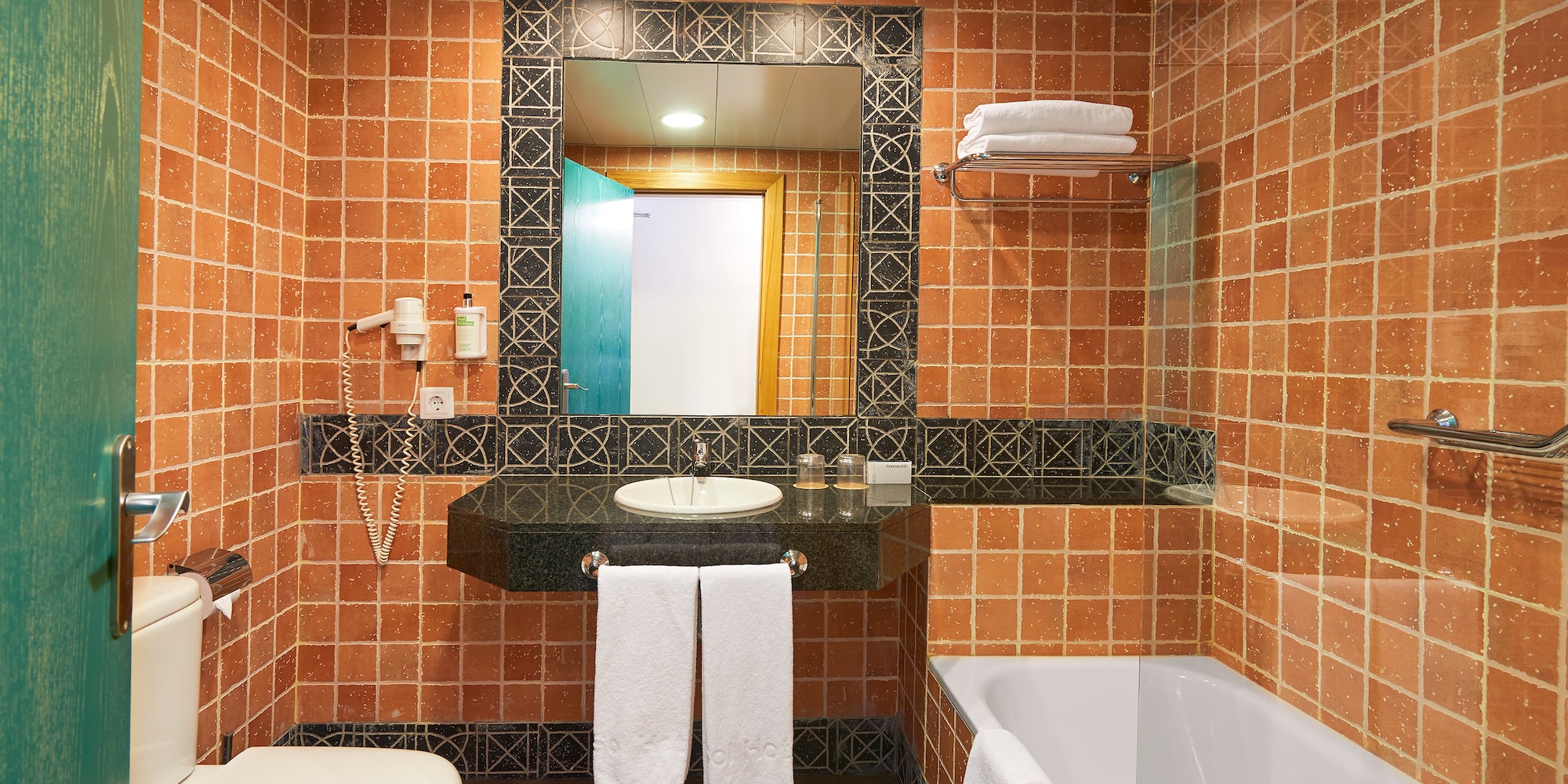 Terracotta tiled hotel bathroom with sink, mirror, toilet, and bathtub.