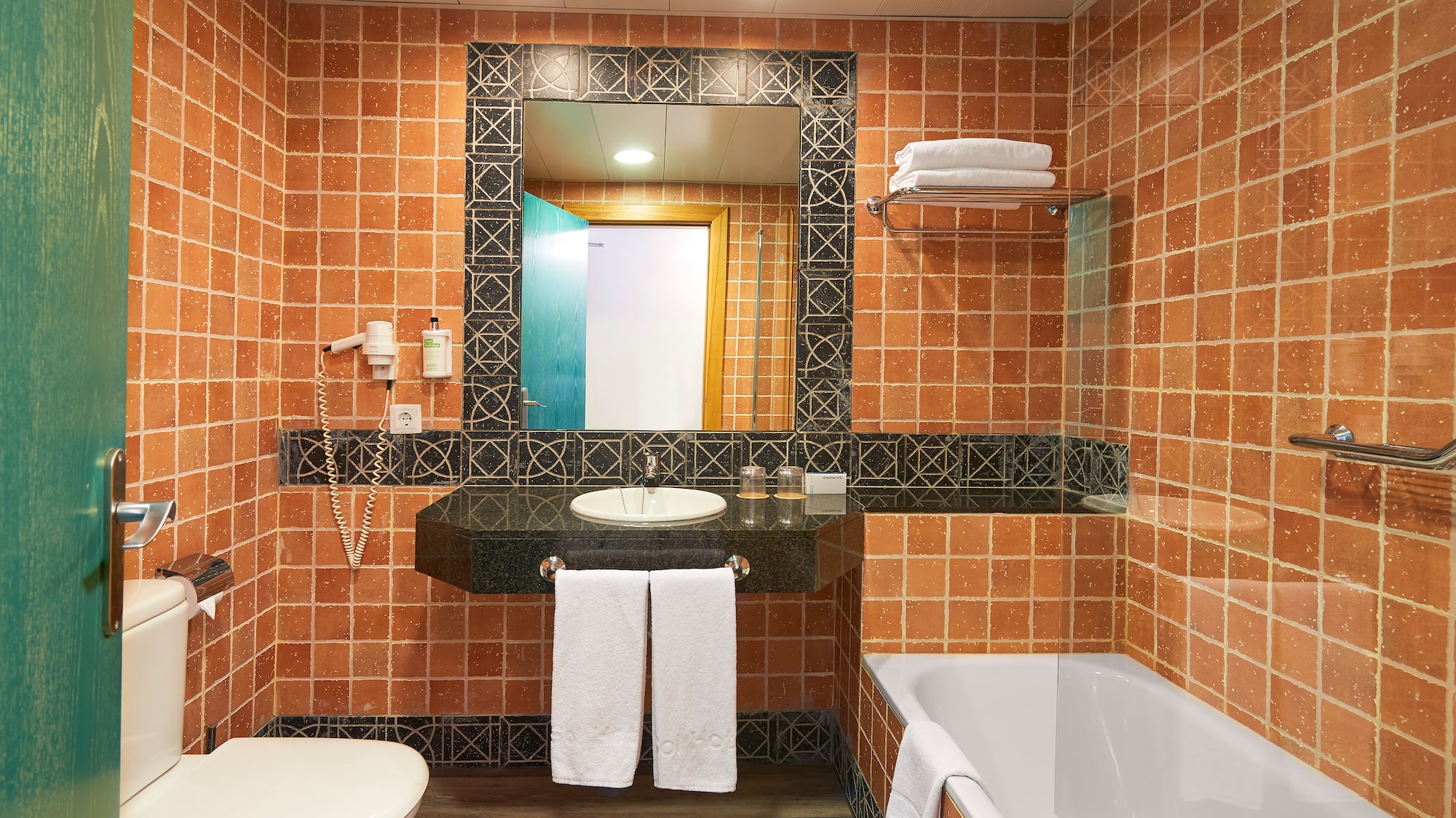 Terracotta tiled hotel bathroom with sink, mirror, toilet, and bathtub.