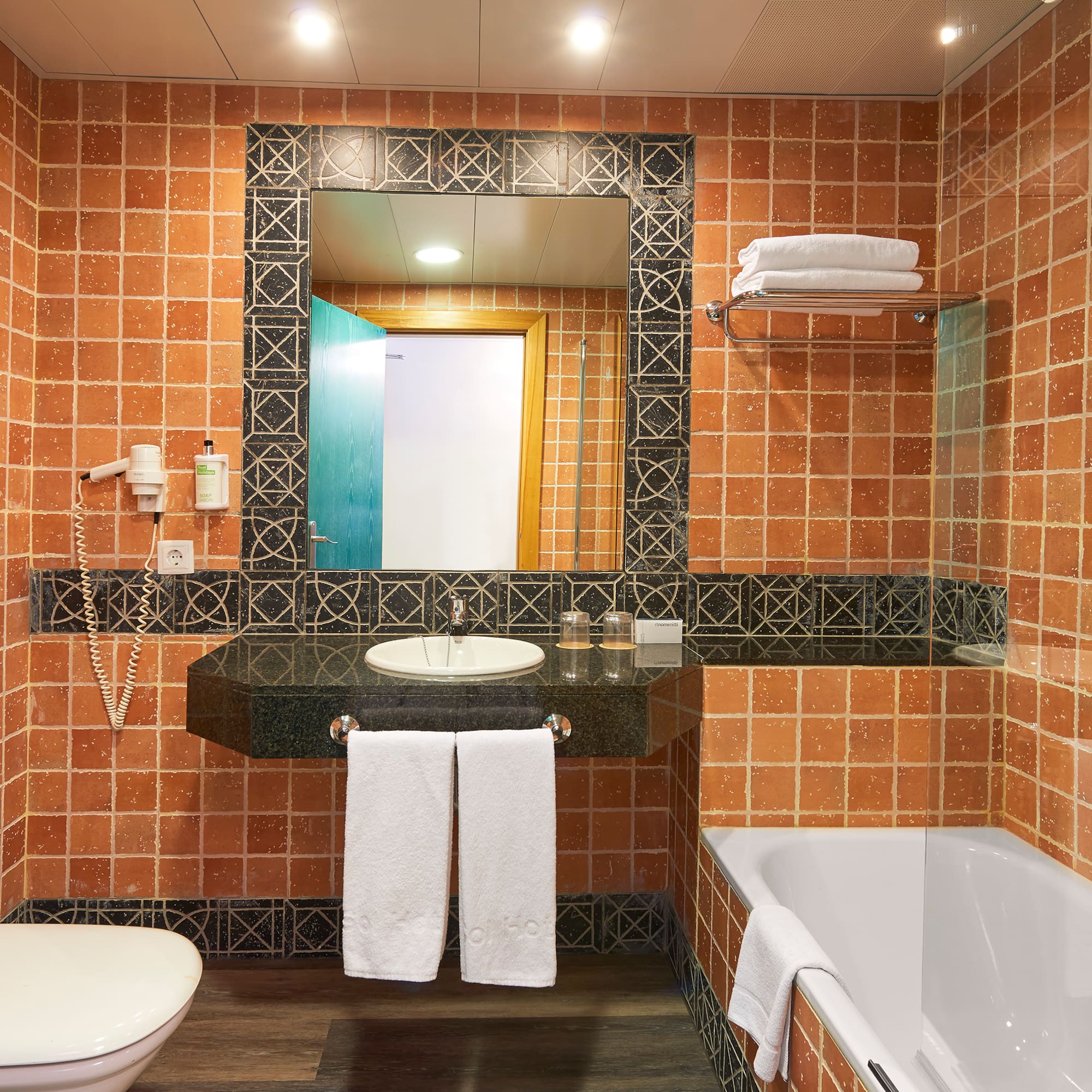 Terracotta tiled hotel bathroom with sink, mirror, toilet, and bathtub.
