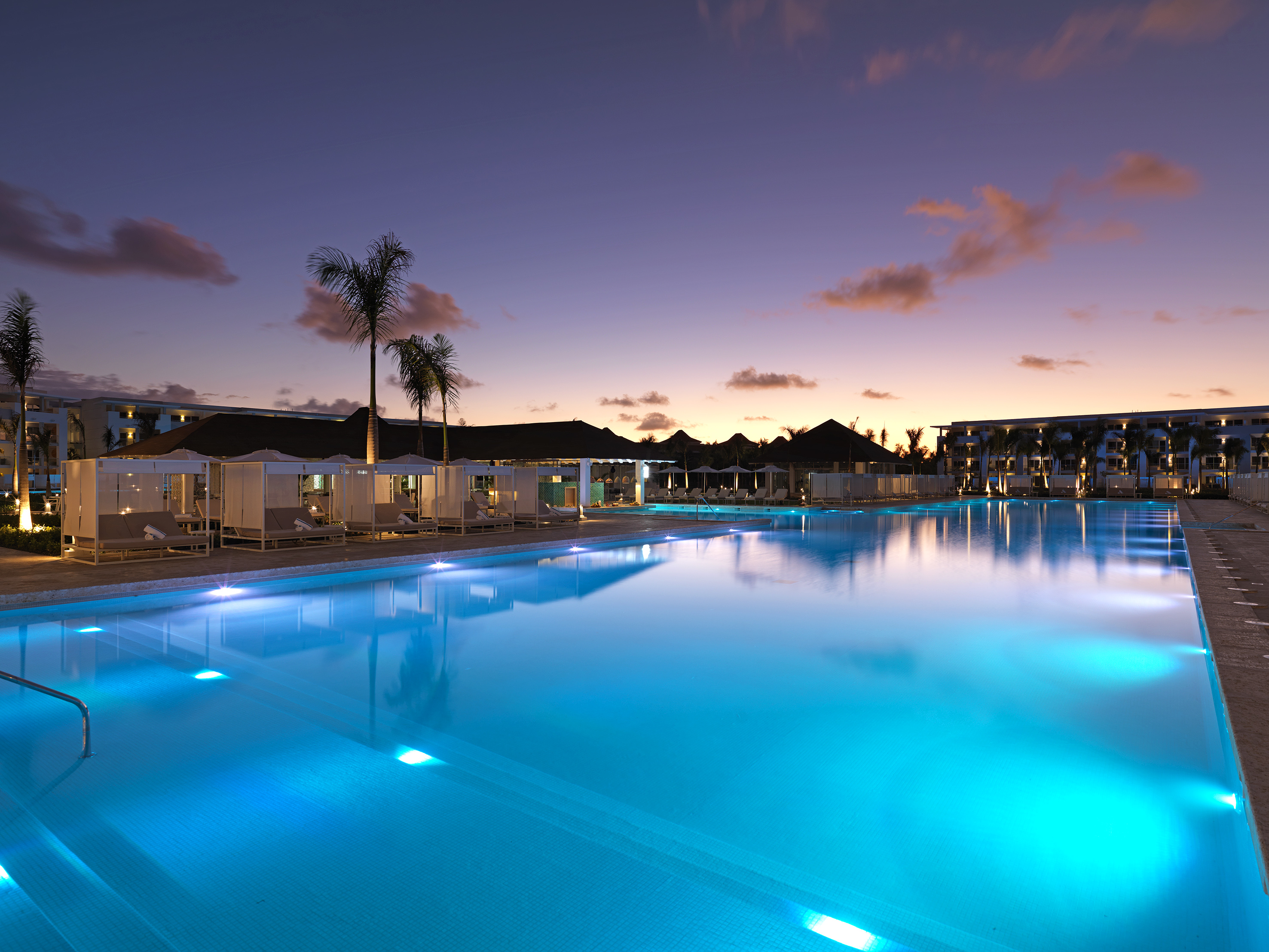Tropical resort pool glowing blue at dusk, with palm trees and cabanas.