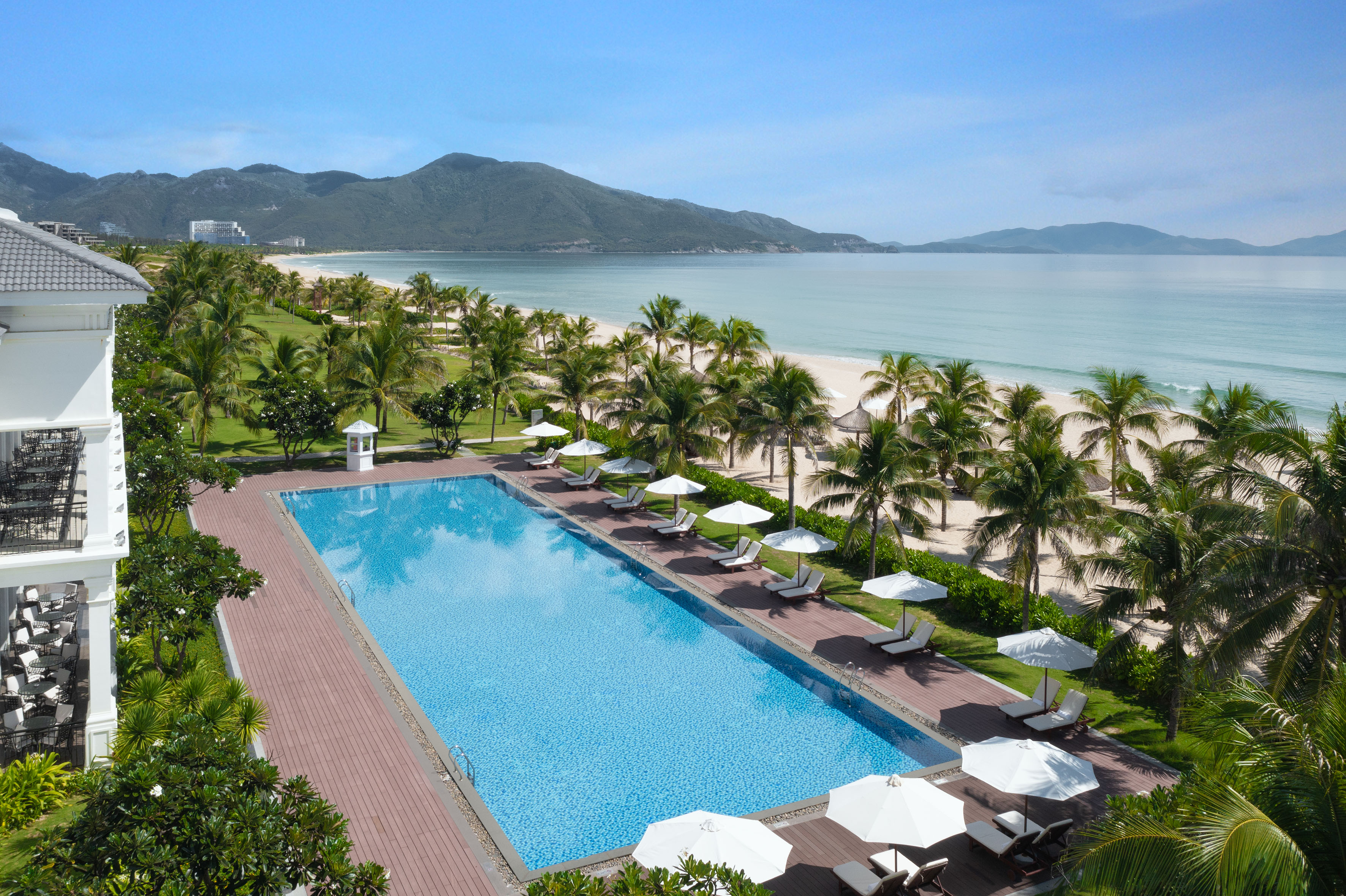 Luxury resort swimming pool overlooking a tropical beach with palm trees and mountains.