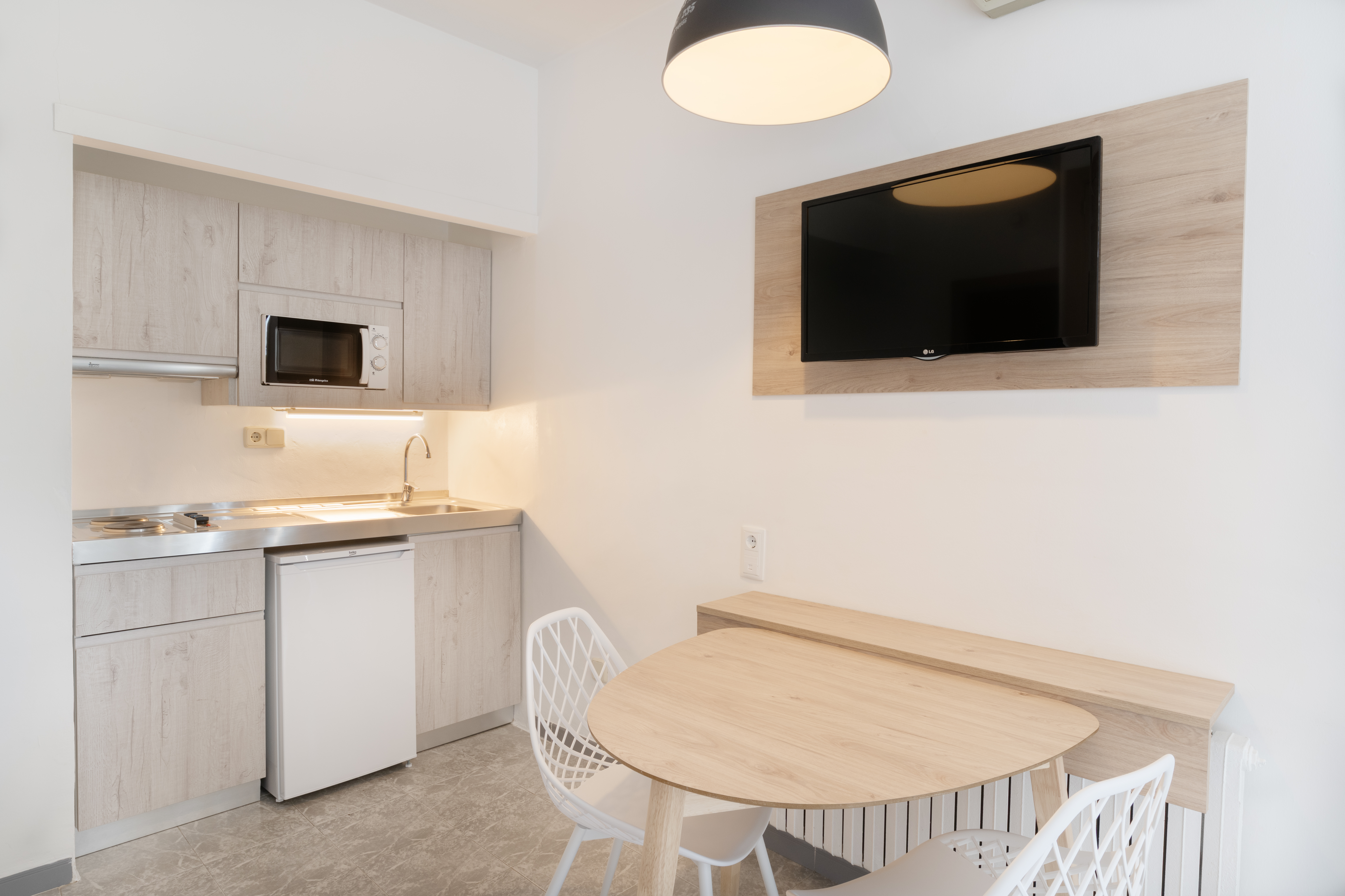 Modern studio kitchenette with light wood cabinets, dining table, and wall TV.