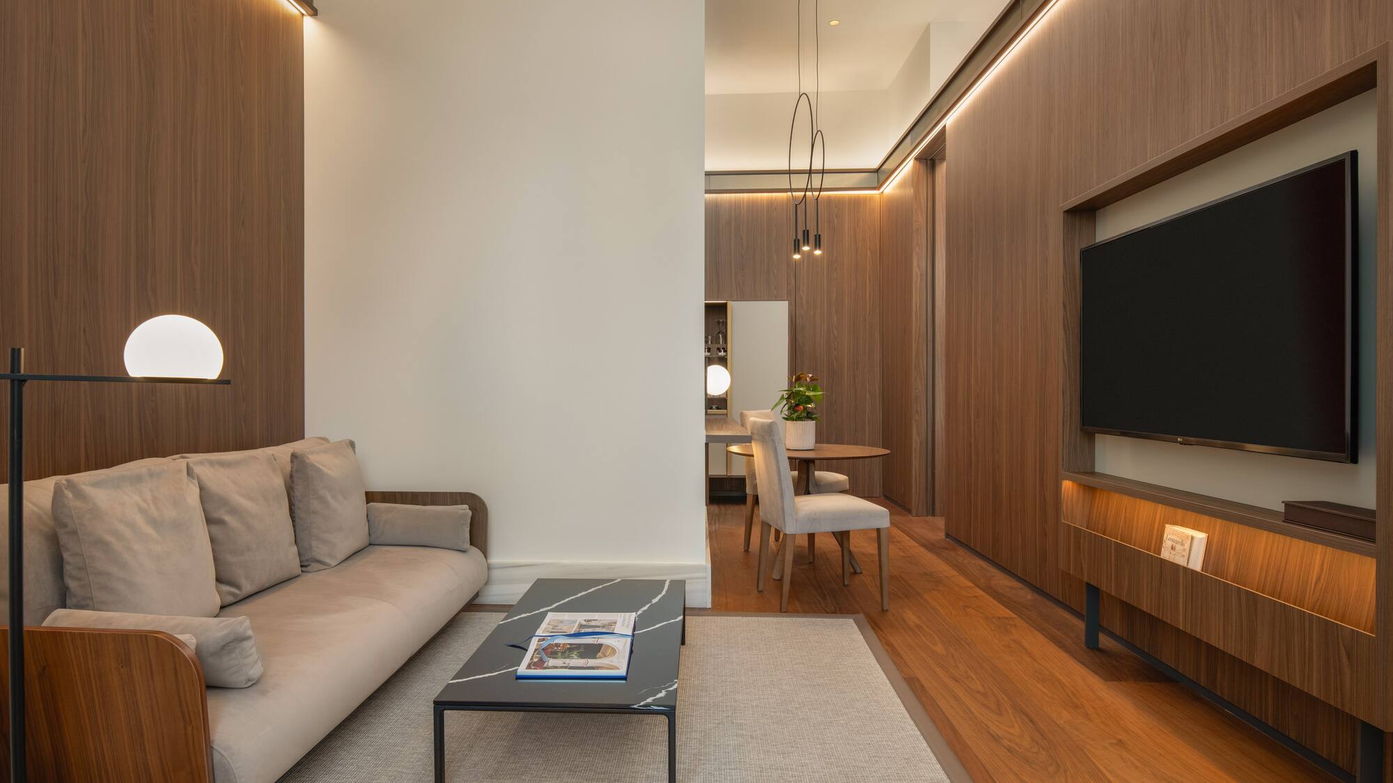 Contemporary living room with wood panels, beige sofa, coffee table, TV, and dining area.