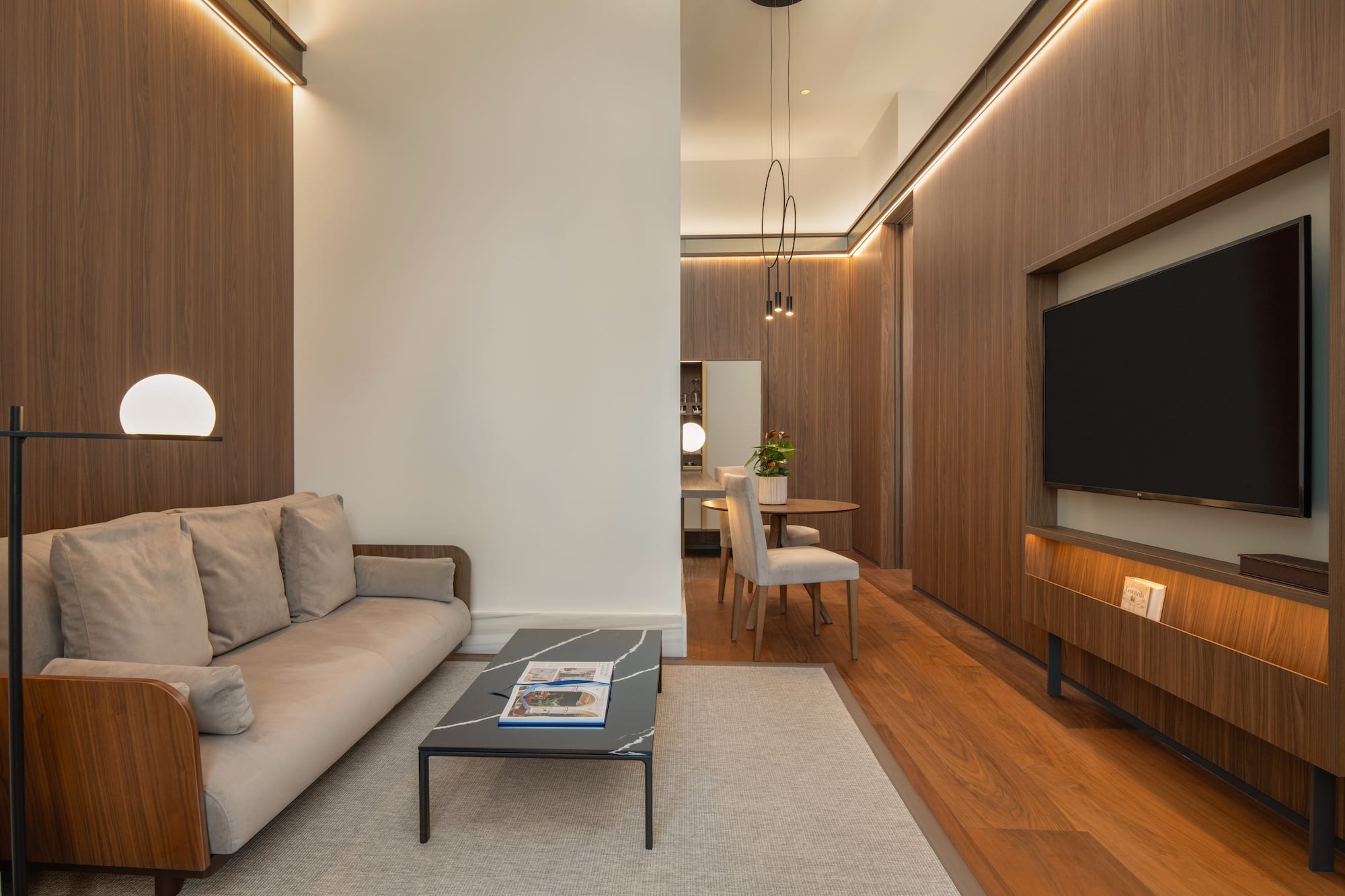 Contemporary living room with wood panels, beige sofa, coffee table, TV, and dining area.