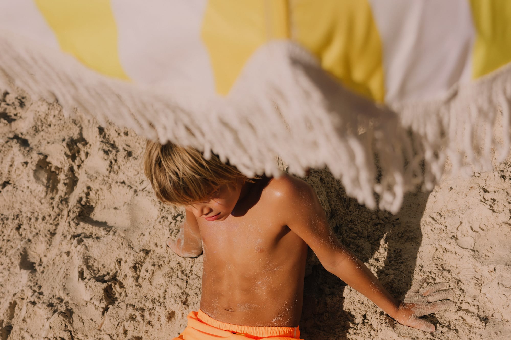 Niño en la arena con shorts naranja bajo sombrilla de rayas amarillas y blancas.