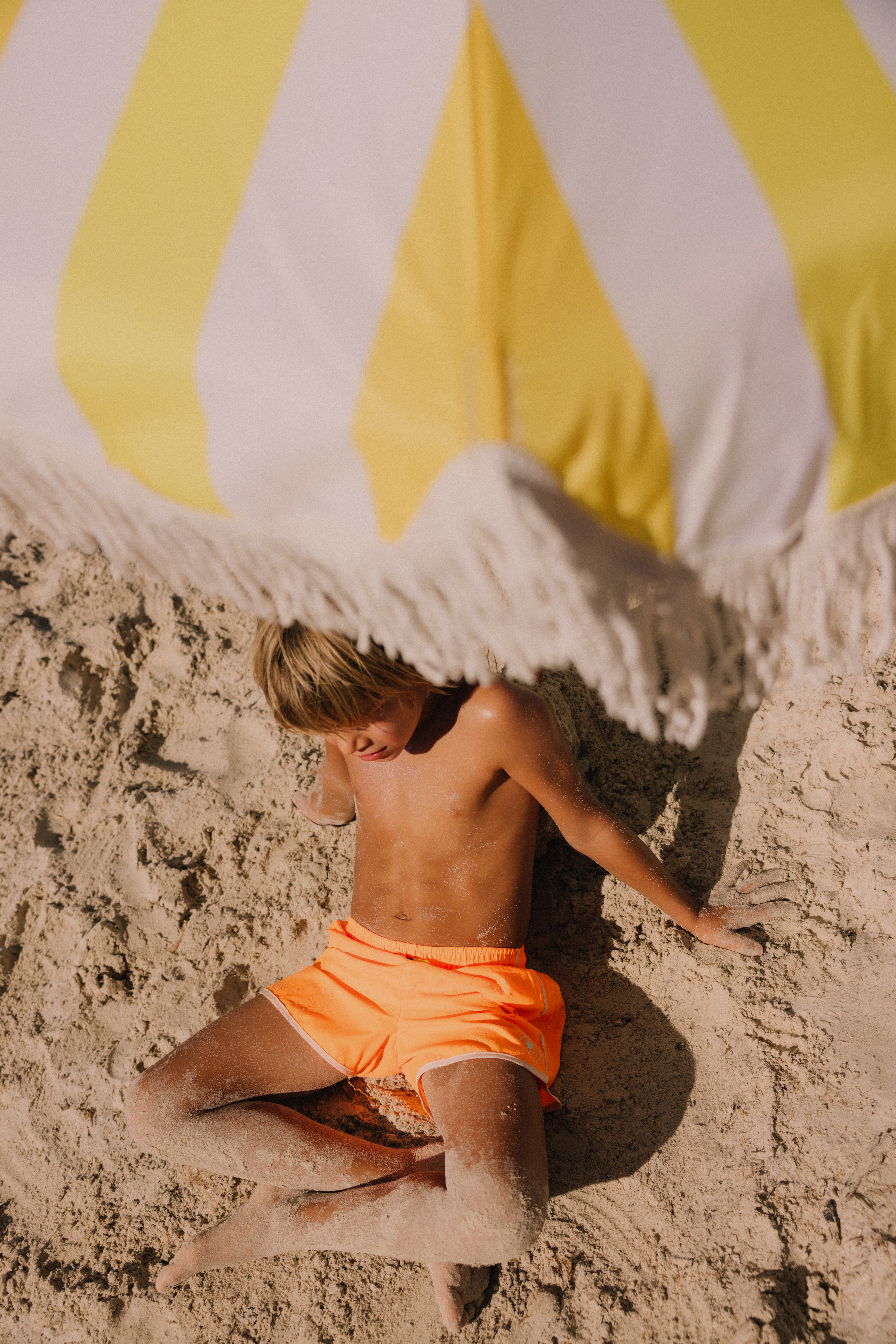 Niño en la arena con shorts naranja bajo sombrilla de rayas amarillas y blancas.