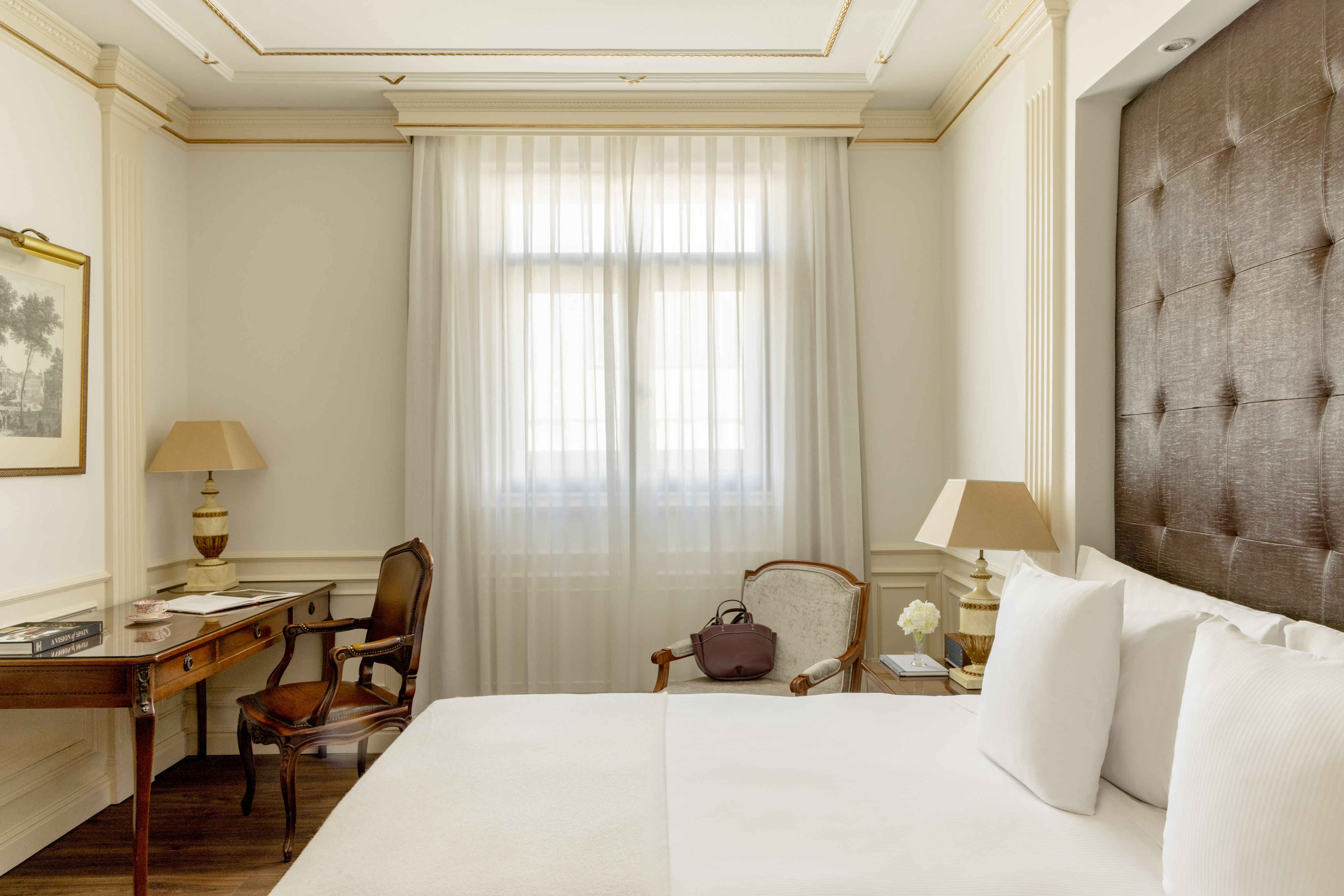 Elegant hotel room with white bed, classic wooden desk, gold crown molding, bright window.