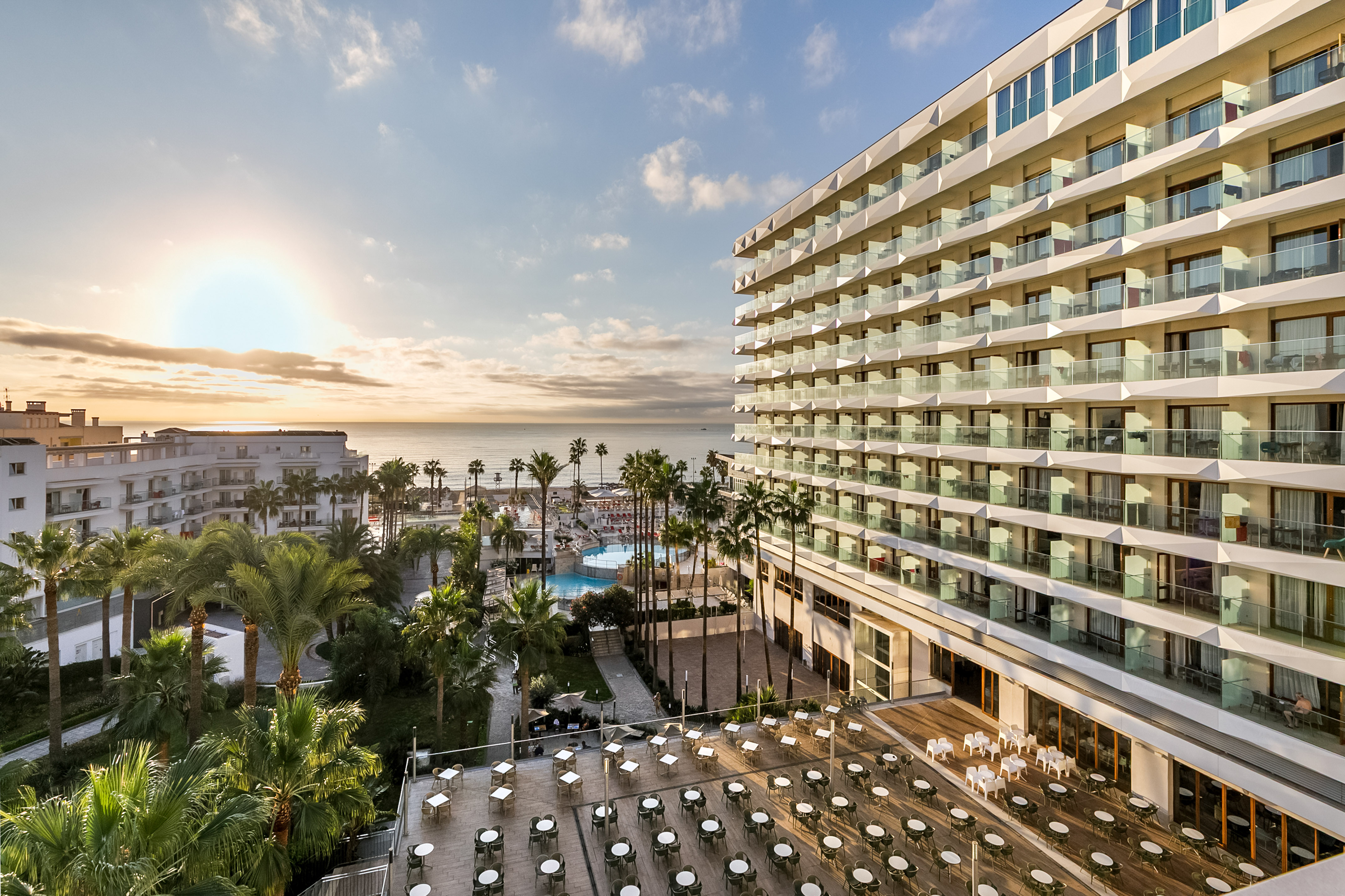 Oceanfront resort with balconies, palm trees, pool, and sunlit outdoor dining.