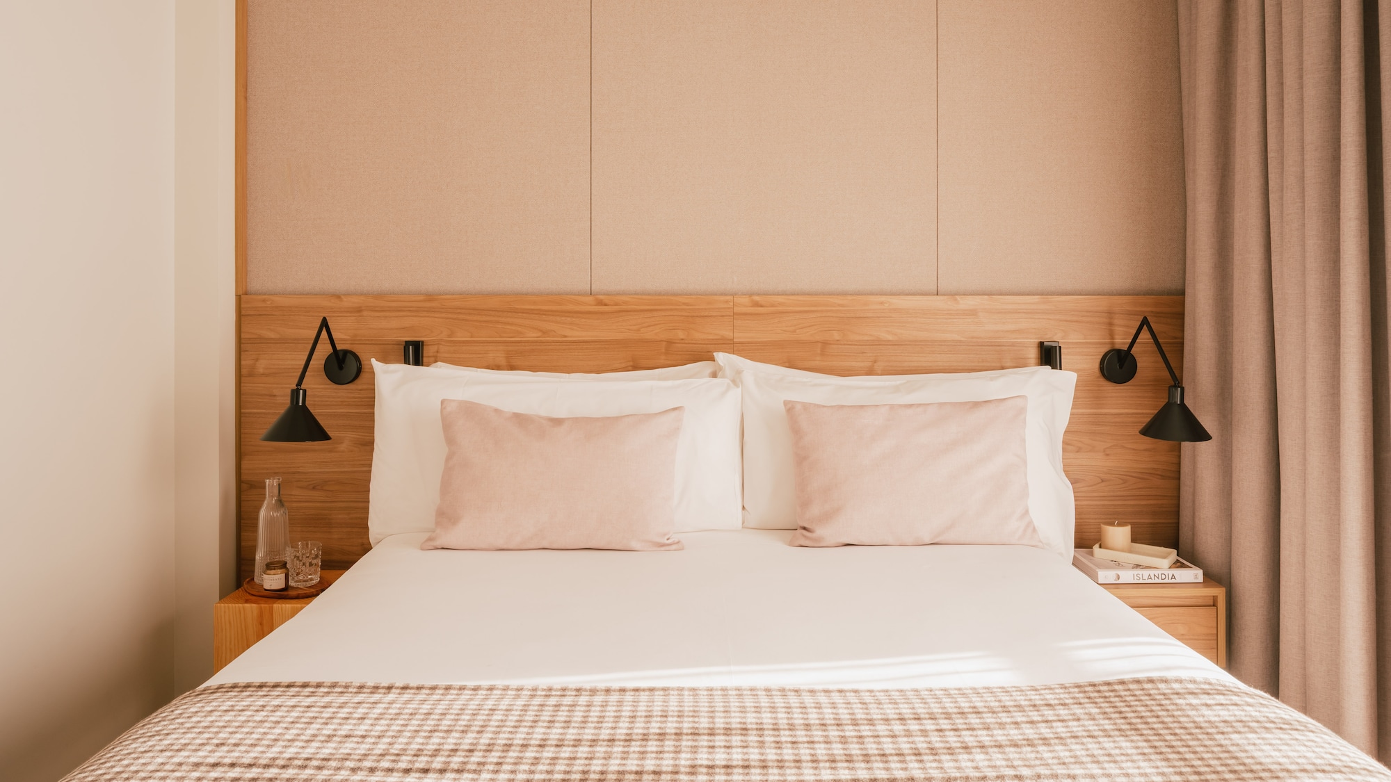 Modern hotel bedroom with wooden headboard, pink pillows, and plaid throw blanket.