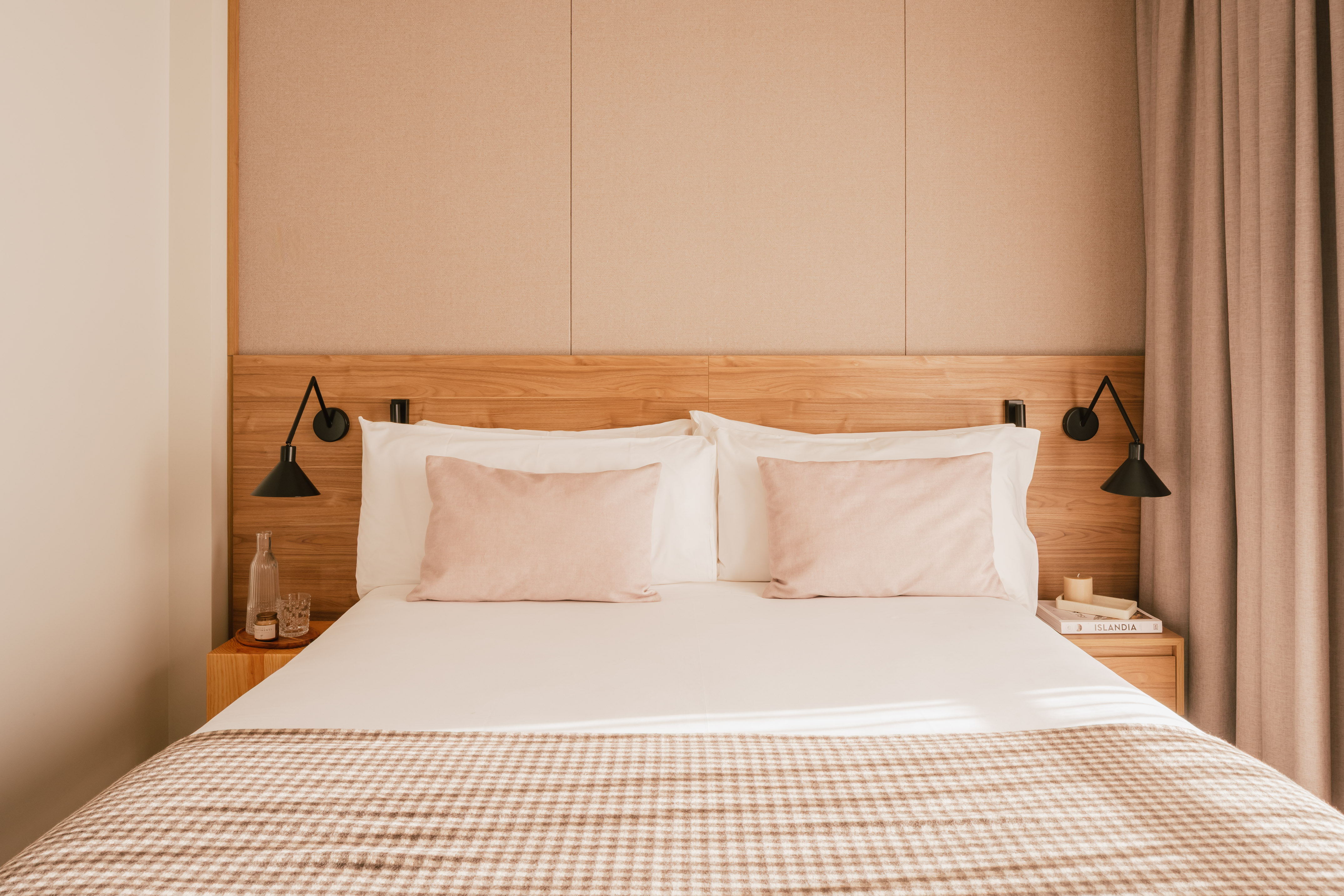 Modern hotel bedroom with wooden headboard, pink pillows, and plaid throw blanket.