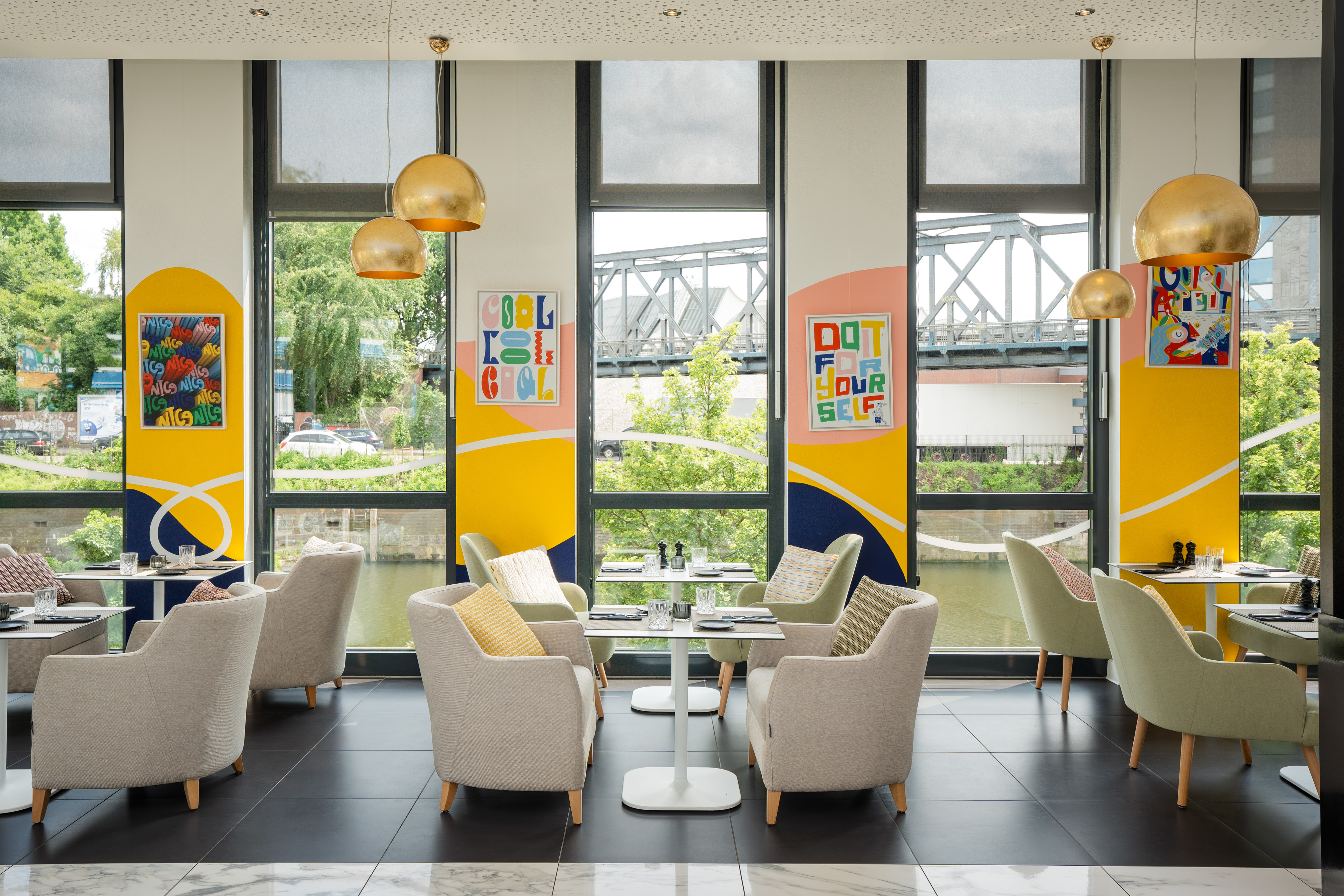 Modern restaurant with colorful art, gold lights, armchairs, and canal view.