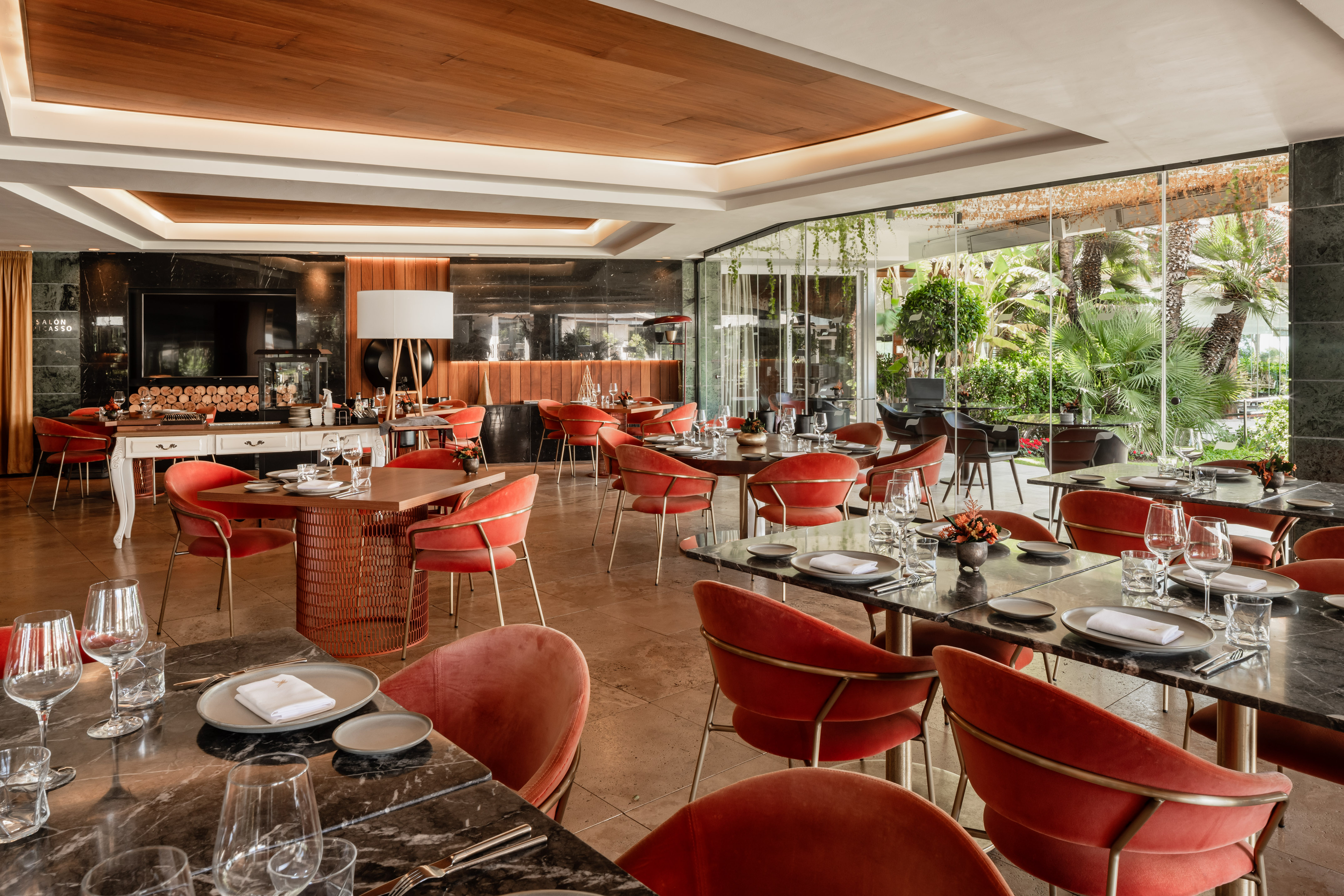 Modern restaurant interior with red velvet chairs, marble tables, and garden view.