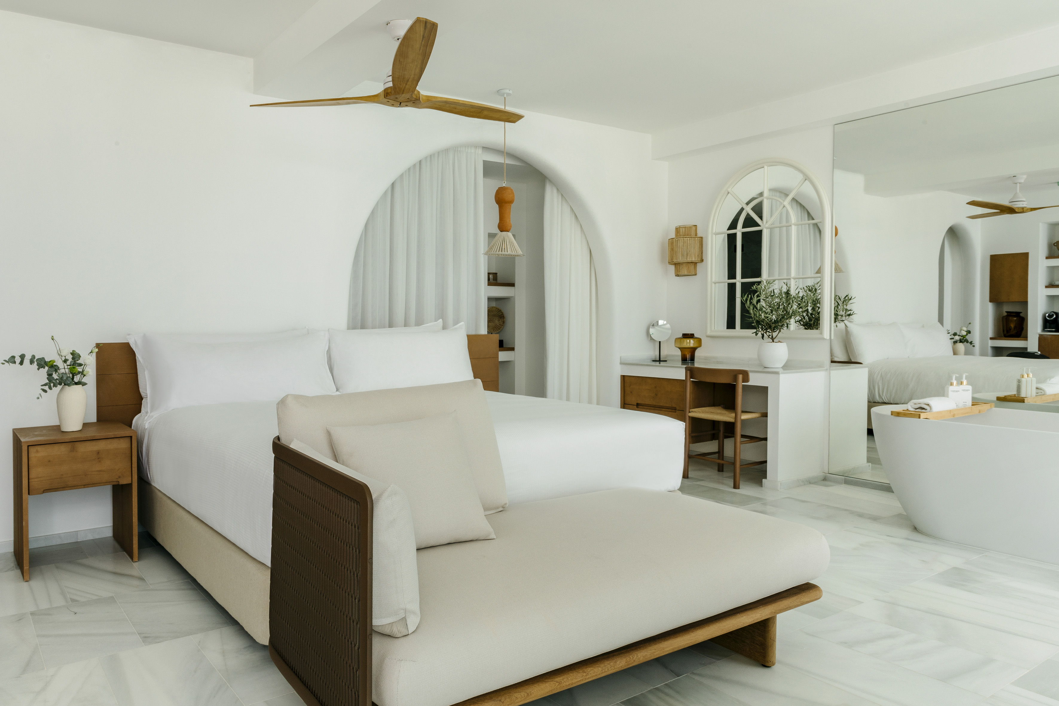 Modern white bedroom with large bed, wood accents, chaise, and bathtub.