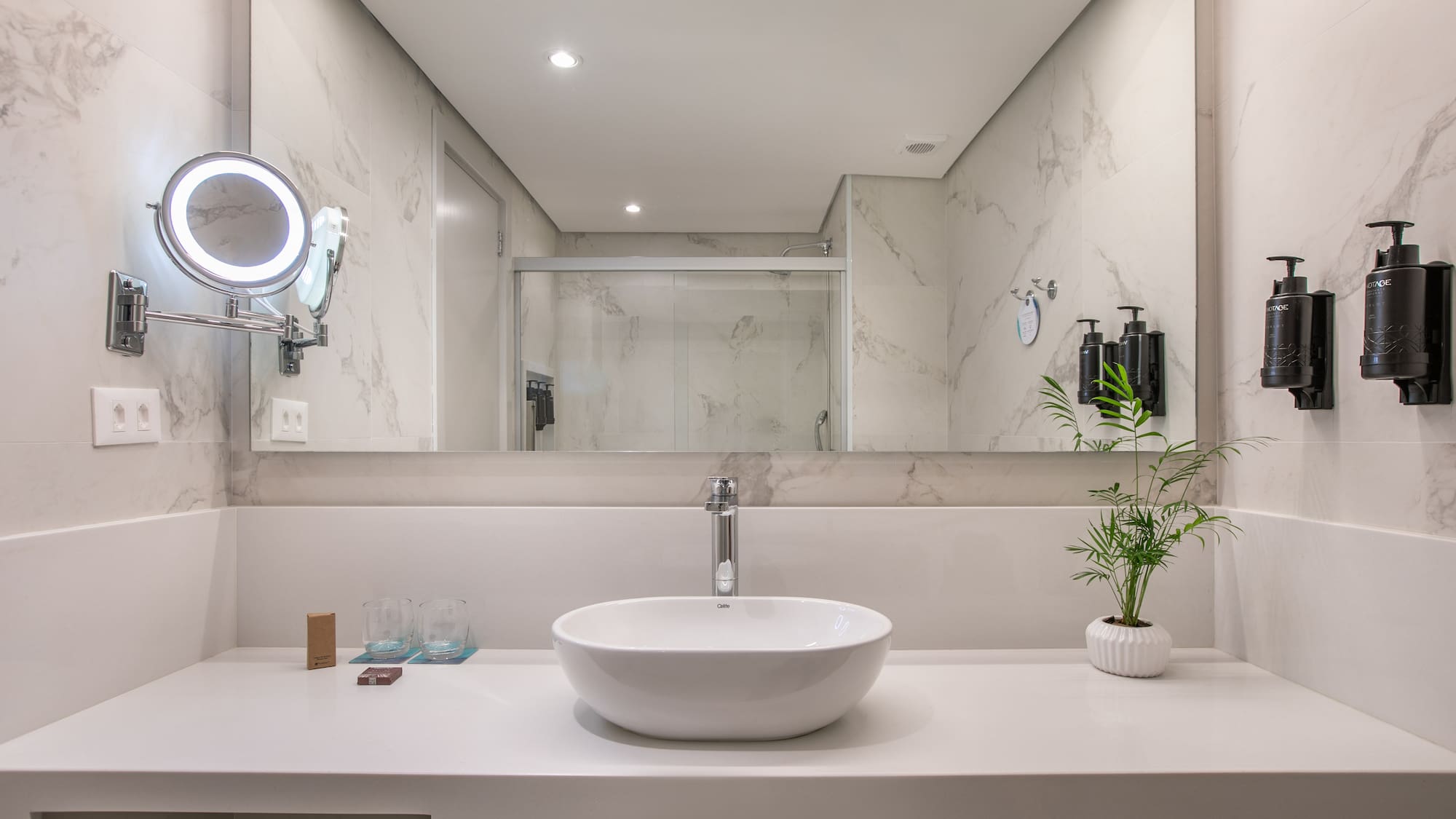 Modern hotel bathroom vanity, white vessel sink, marble walls, lighted mirror.