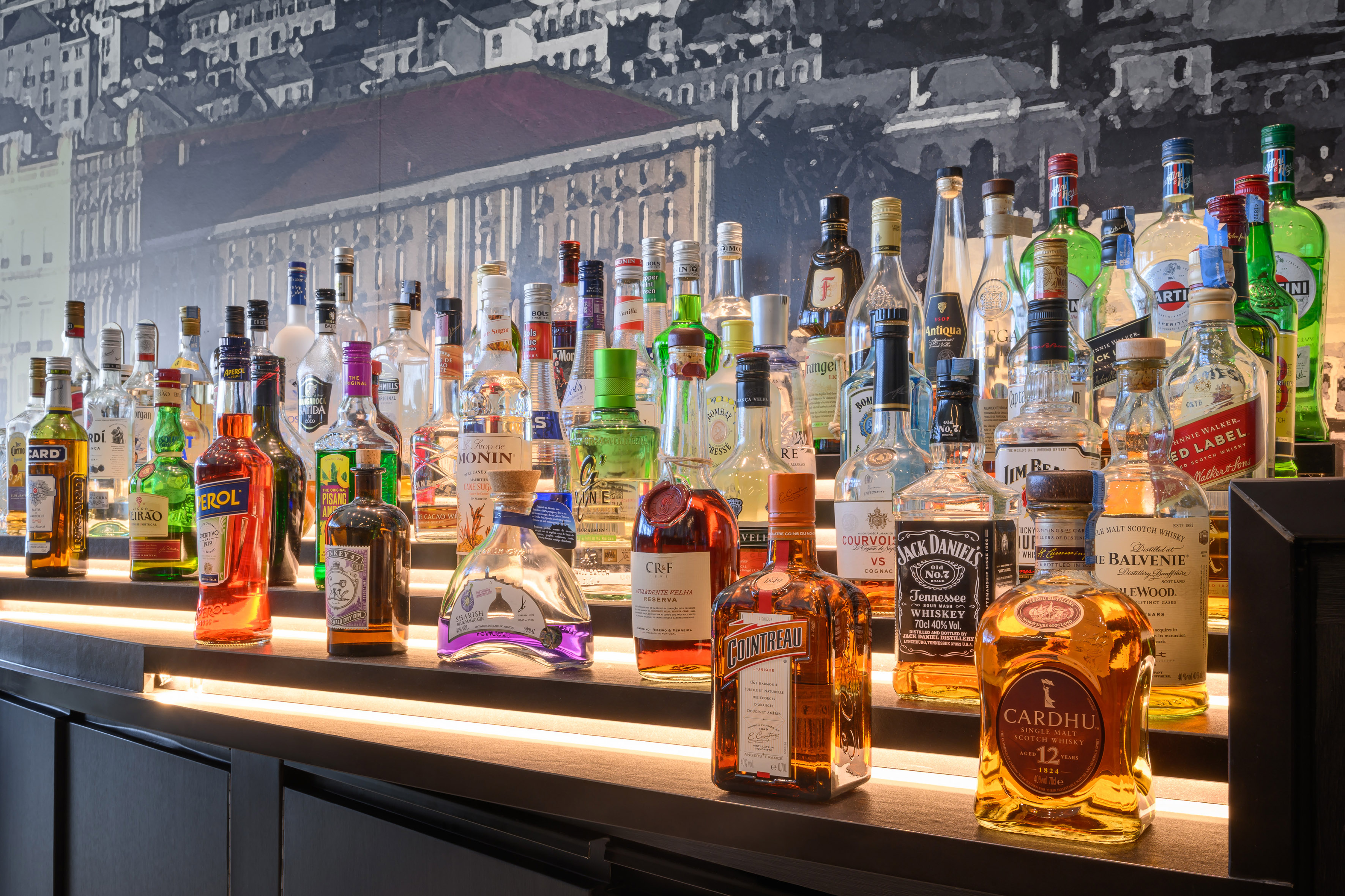 Assorted liquor bottles on illuminated bar shelves, city mural background.