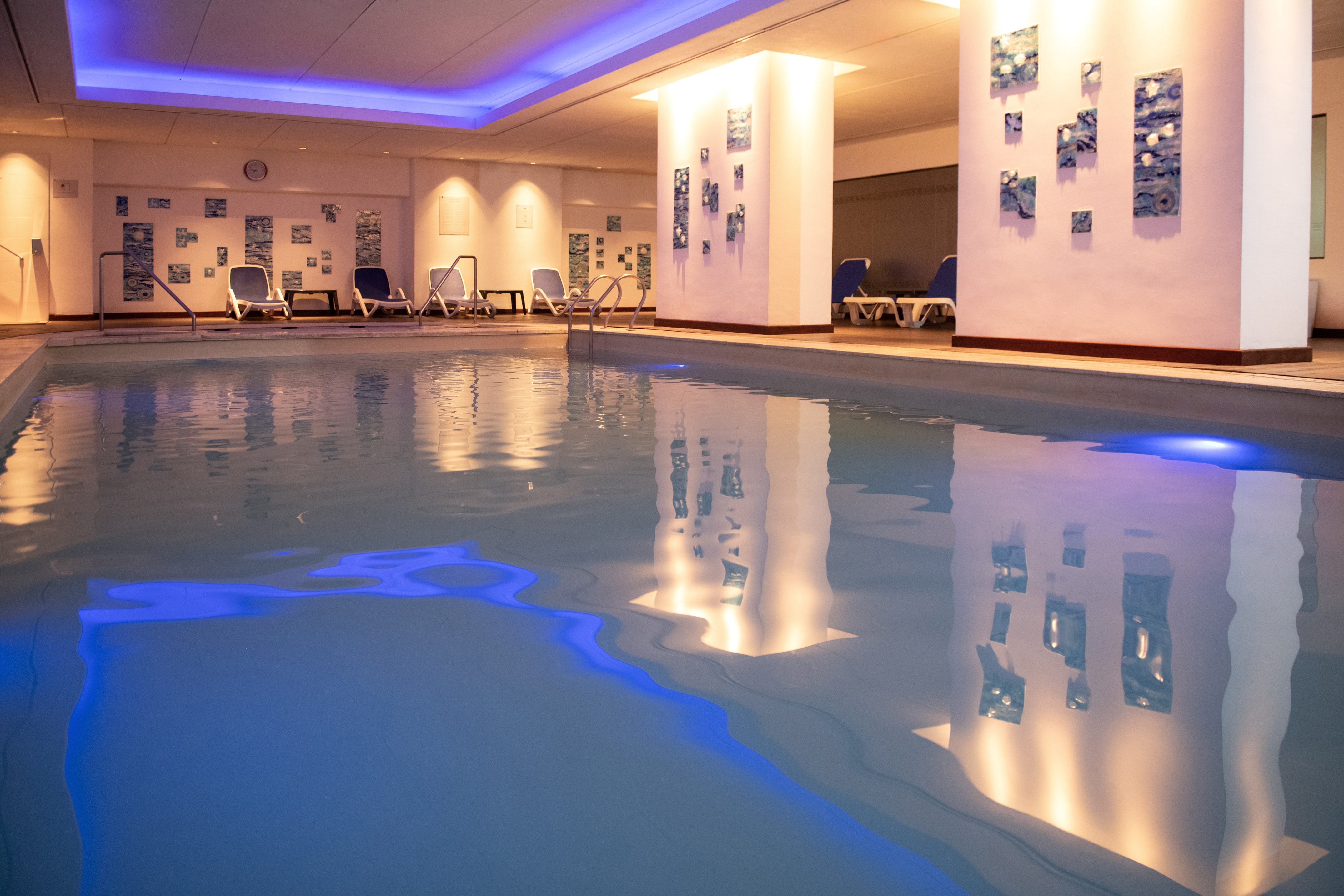 Indoor hotel pool with blue lighting, lounge chairs, and wall-mounted art tiles.