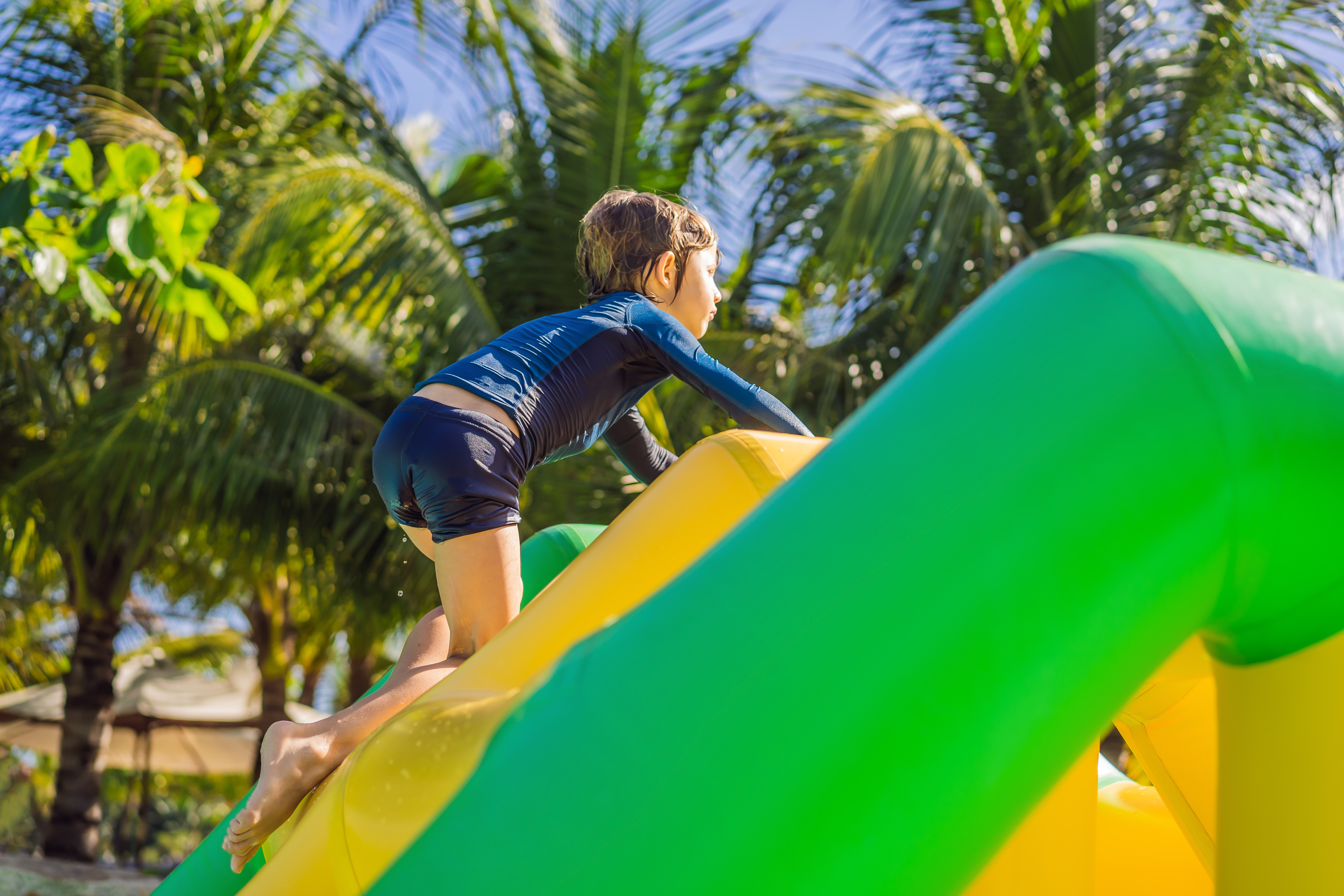 Niño subiendo inflable acuático amarillo y verde, bajo palmeras.