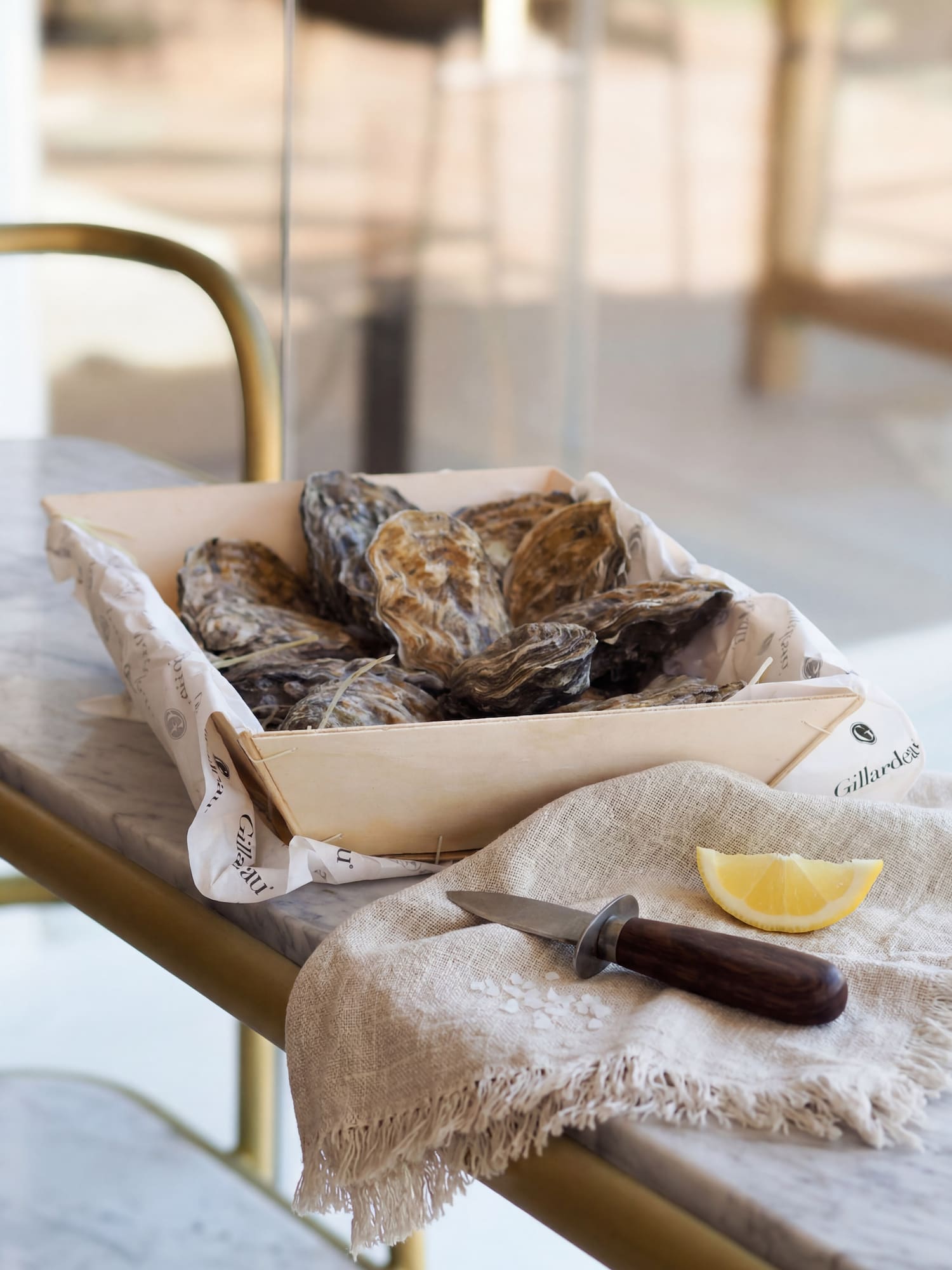 Fresh Gillardeau oysters in a wooden tray with shucking knife and lemon on marble.