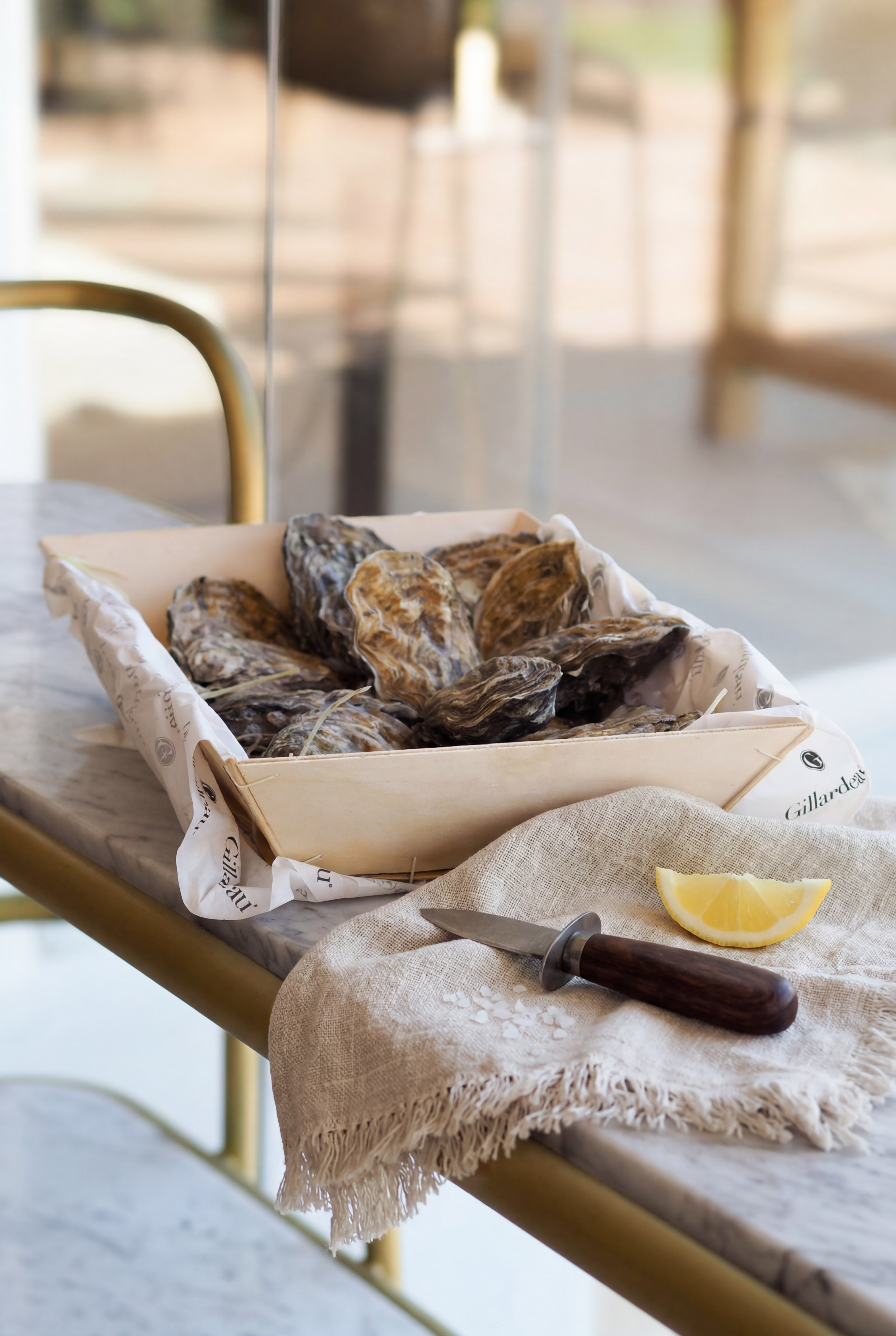 Fresh Gillardeau oysters in a wooden tray with shucking knife and lemon on marble.