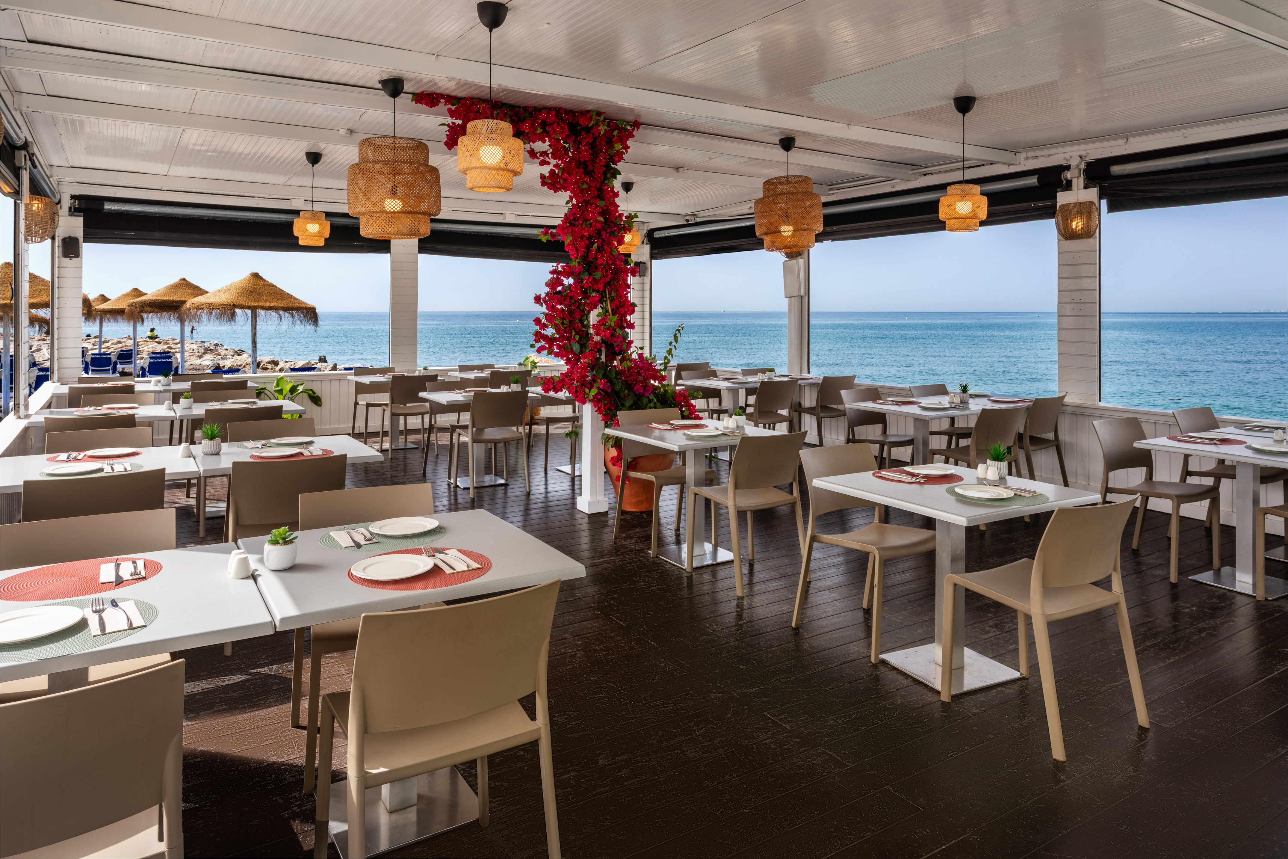 Beachfront restaurant with sea views, set tables, and red floral decor.