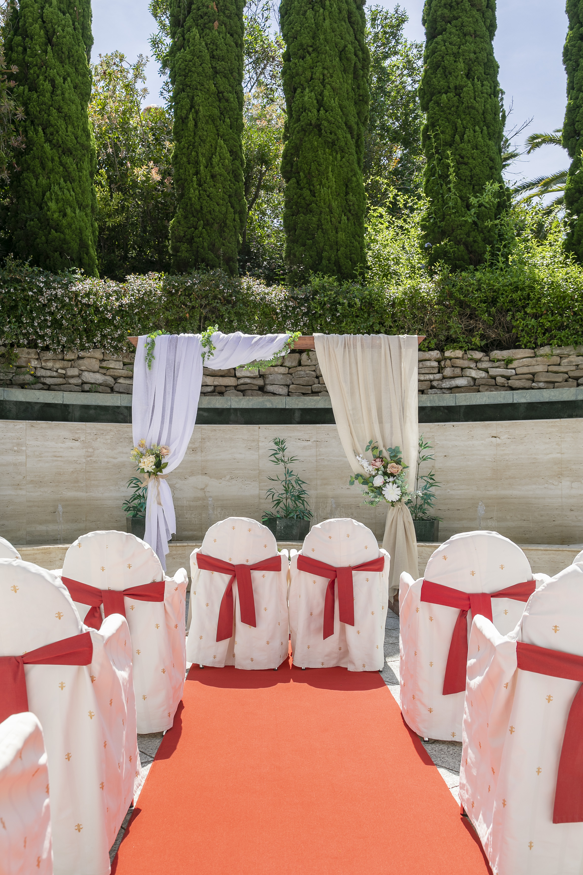 Garden wedding aisle with red carpet, white chairs, and draped floral arch.