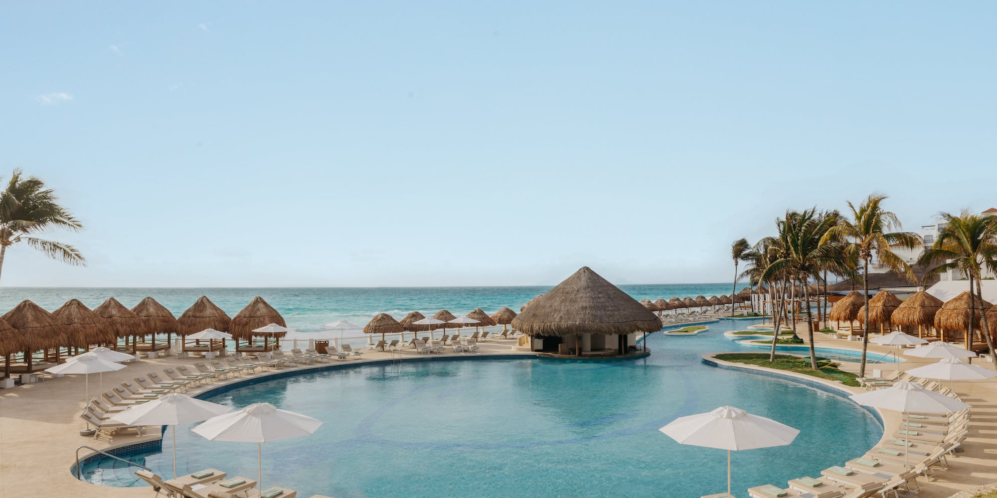 Resort pool with swim-up bar, beach, palapas, and palm trees.