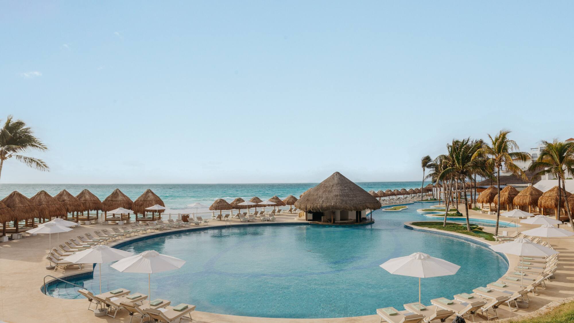 Resort pool with swim-up bar, beach, palapas, and palm trees.