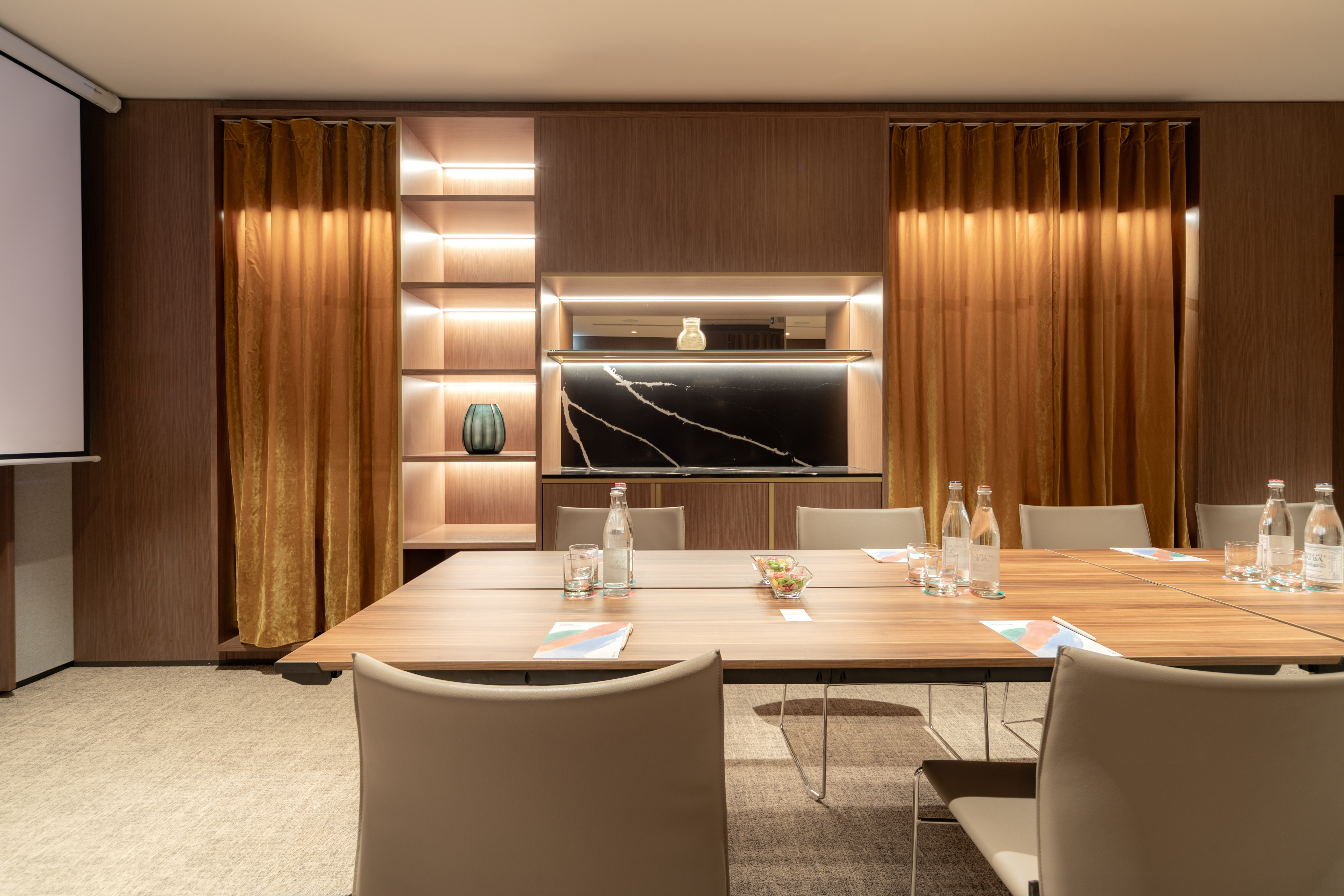 Professional meeting room with wooden table, screen, and elegant lighted shelving.