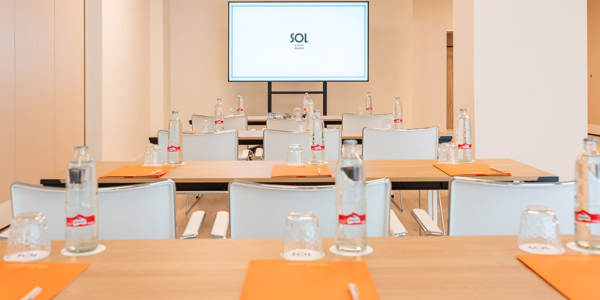 Modern conference room with tables, white chairs, bottled water, and large screen.
