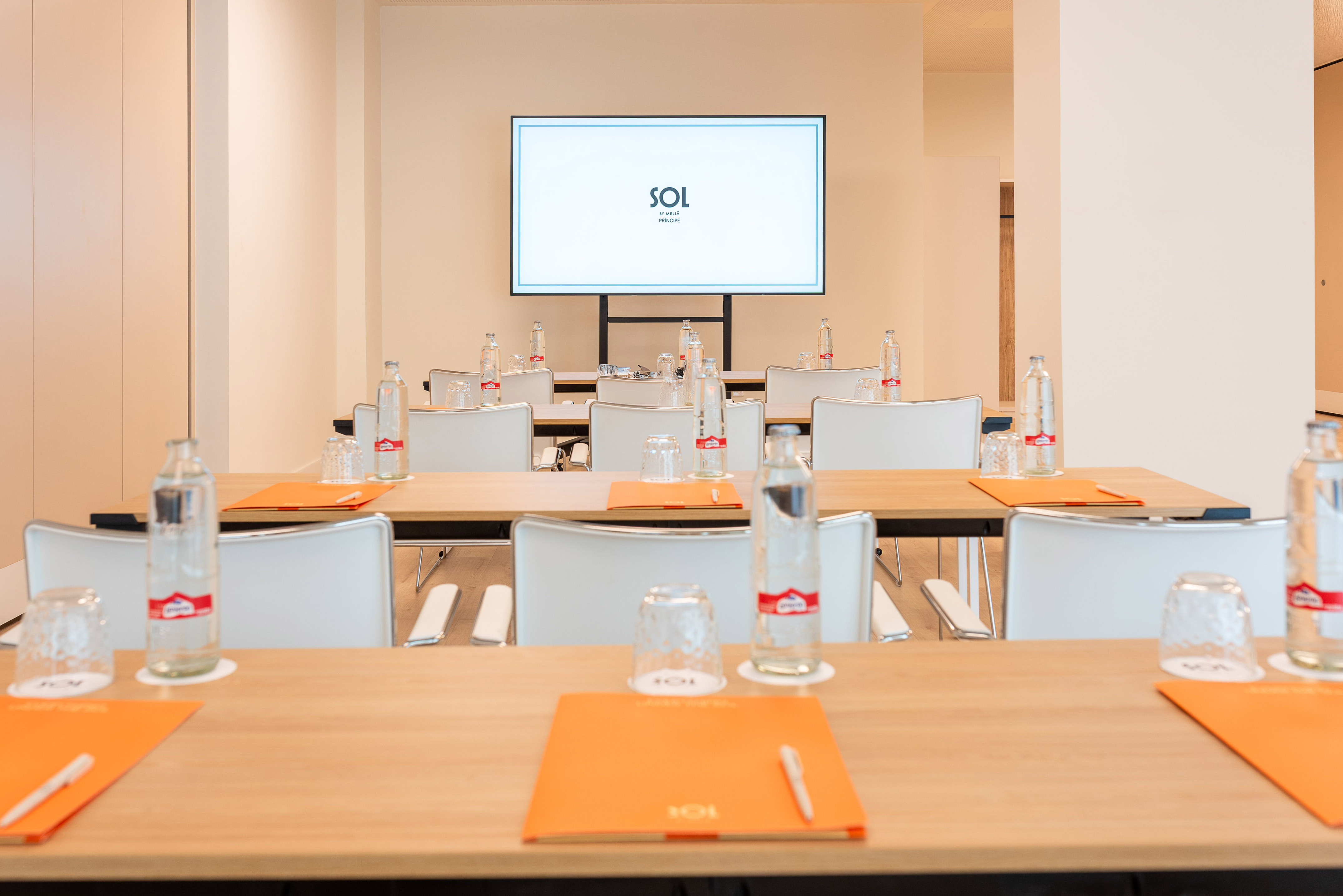 Modern conference room with tables, white chairs, bottled water, and large screen.
