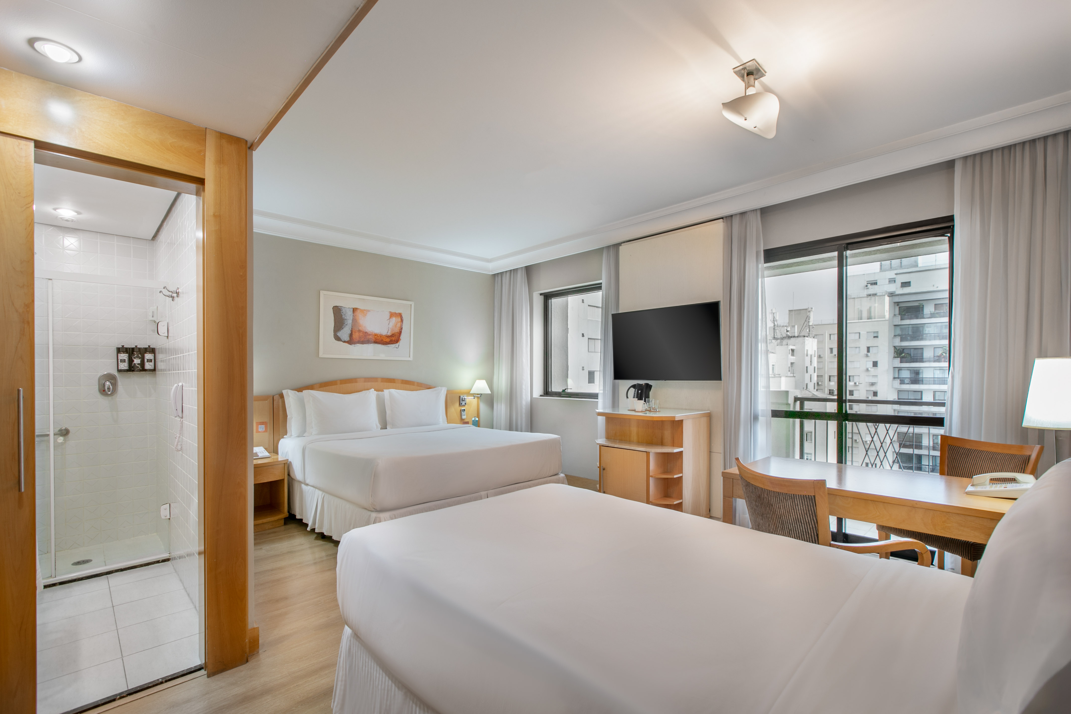 Modern hotel room with two beds, city view, work desk, and shower.