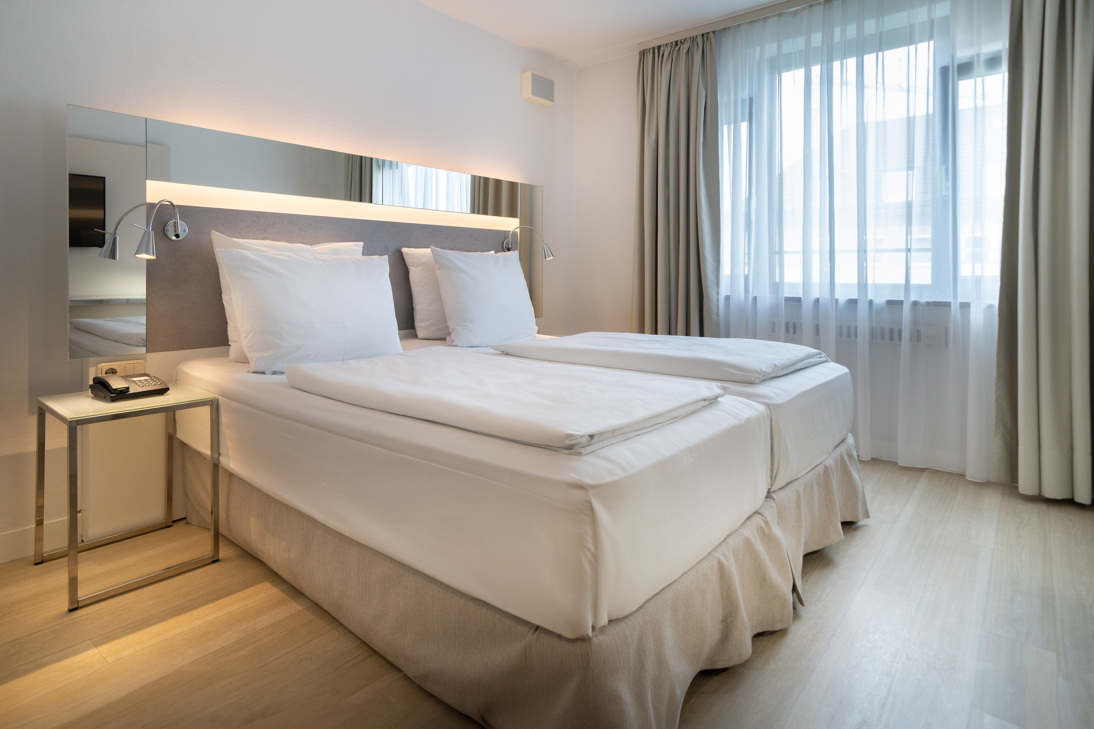 Modern hotel room with twin beds, white bedding, mirrored headboard, and window.