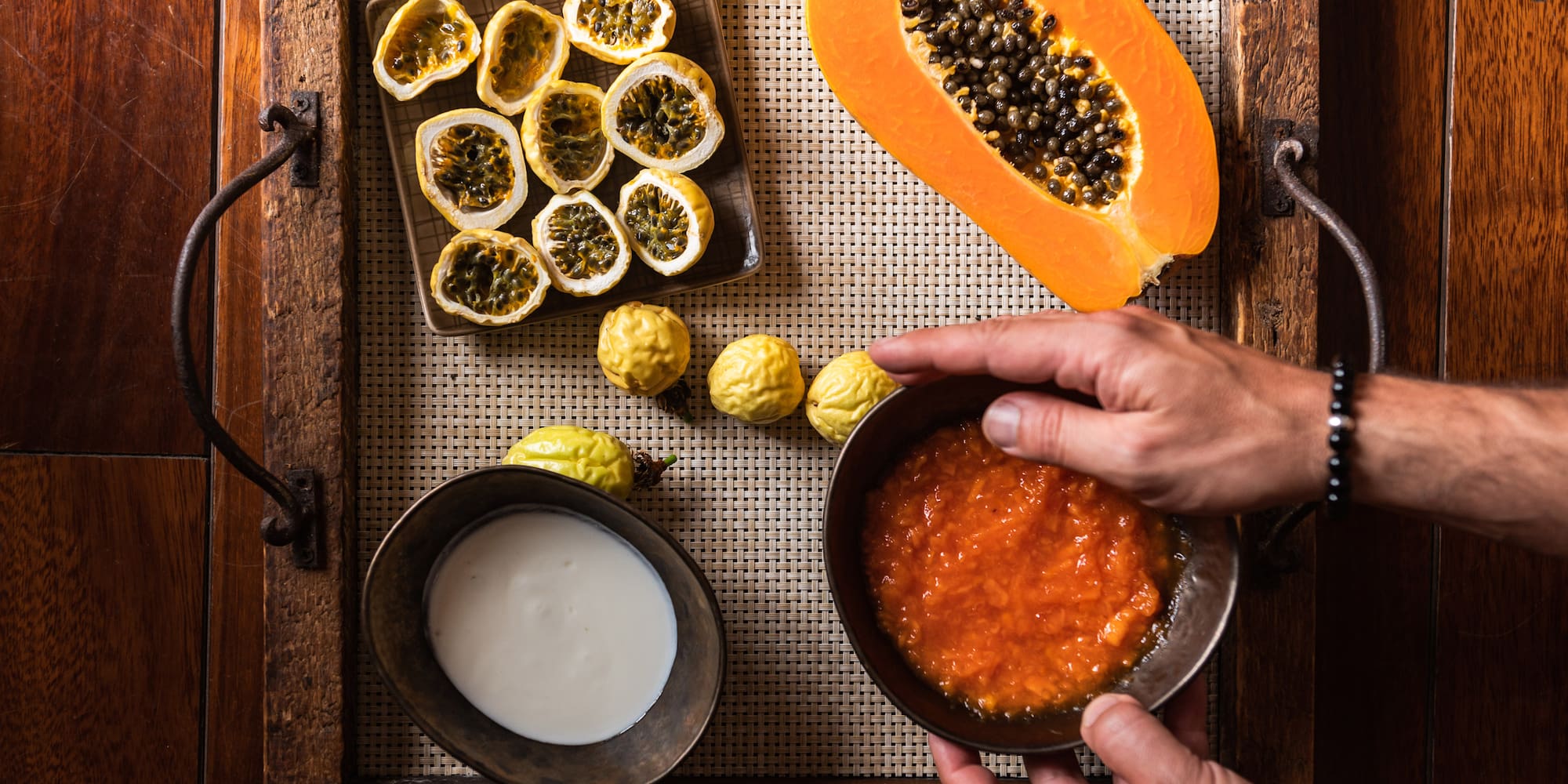 Hands holding papaya pulp bowl; fresh papaya, passion fruits on rustic tray.