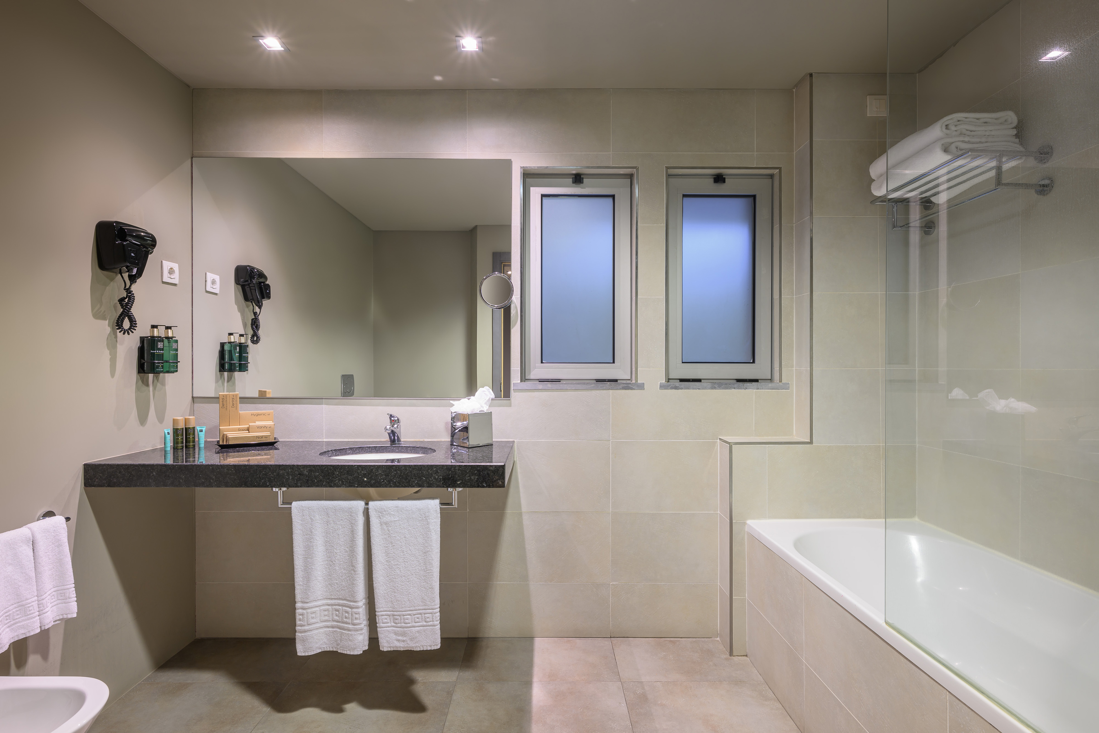 Modern hotel bathroom with vanity, mirror, bathtub, and frosted windows.