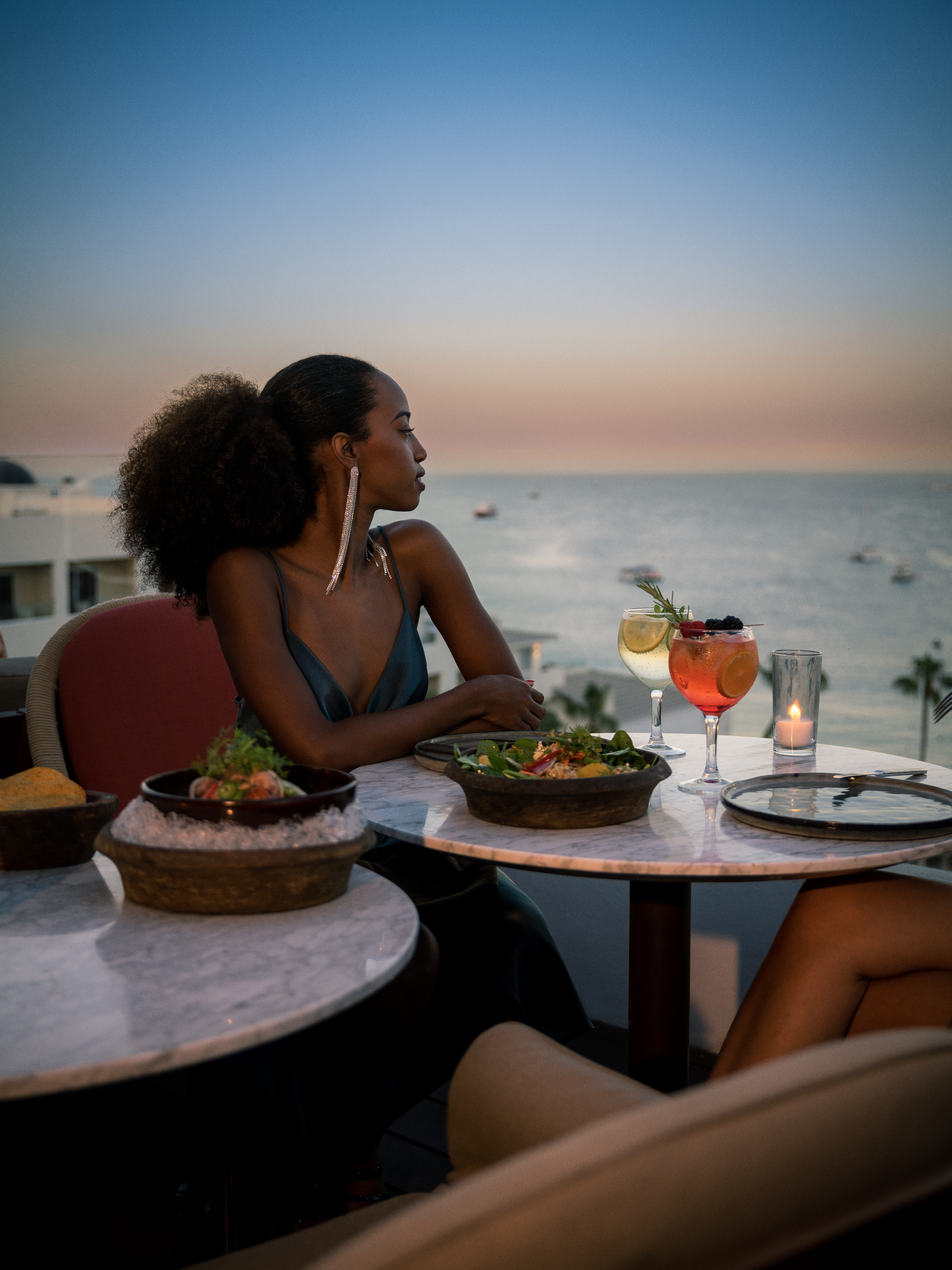 Woman enjoying rooftop dinner with ocean view, cocktails, and sunset.