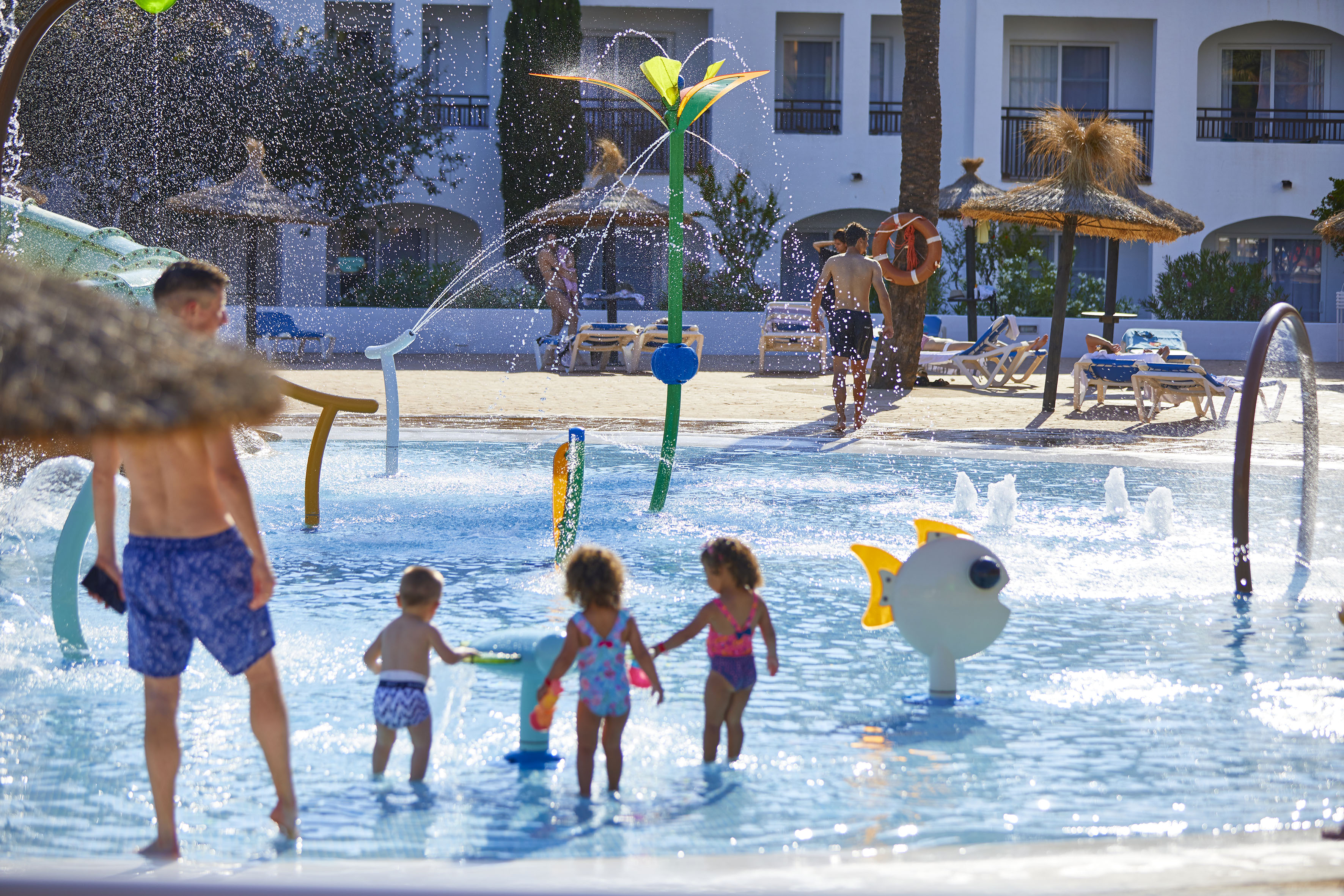 Families play in a vibrant hotel water park splash pad with children and sprinklers.