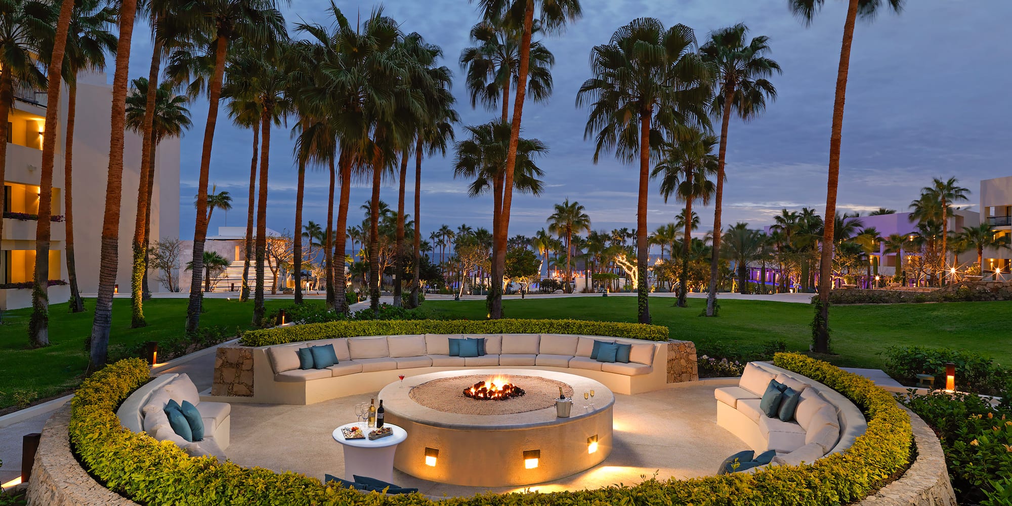 Outdoor resort fire pit lounge with curved seating, palm trees, and evening. - Paradisus Los Cabos - Adults Only, Los Cabos